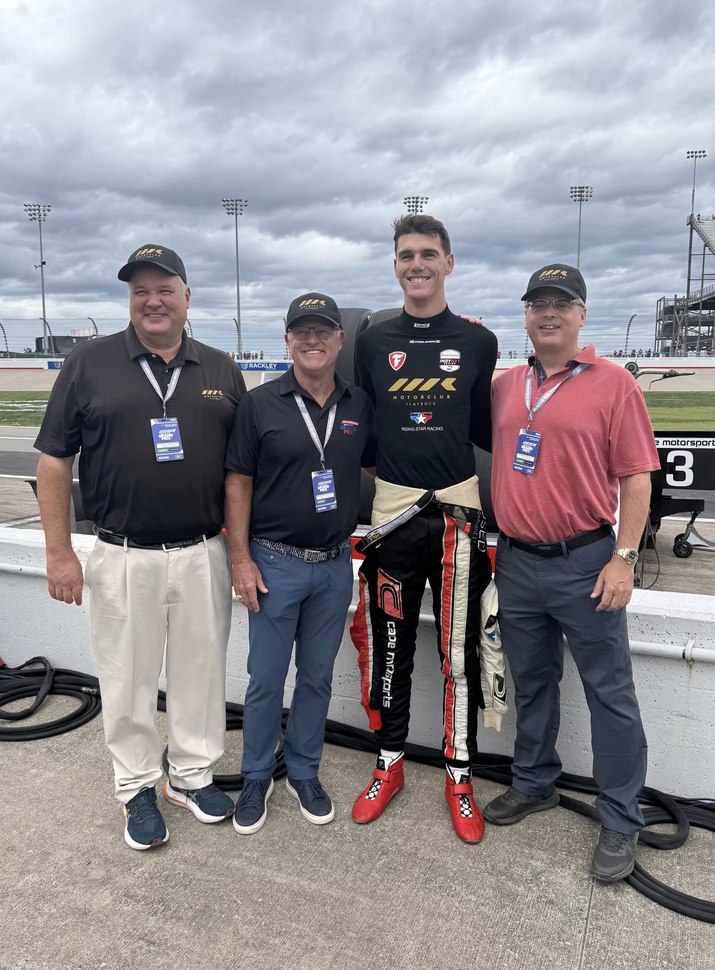 Michael d'Orlando and team on a race track fence, one in racing suit, three in casual clothing, four light poles, and cloudy sky in background.