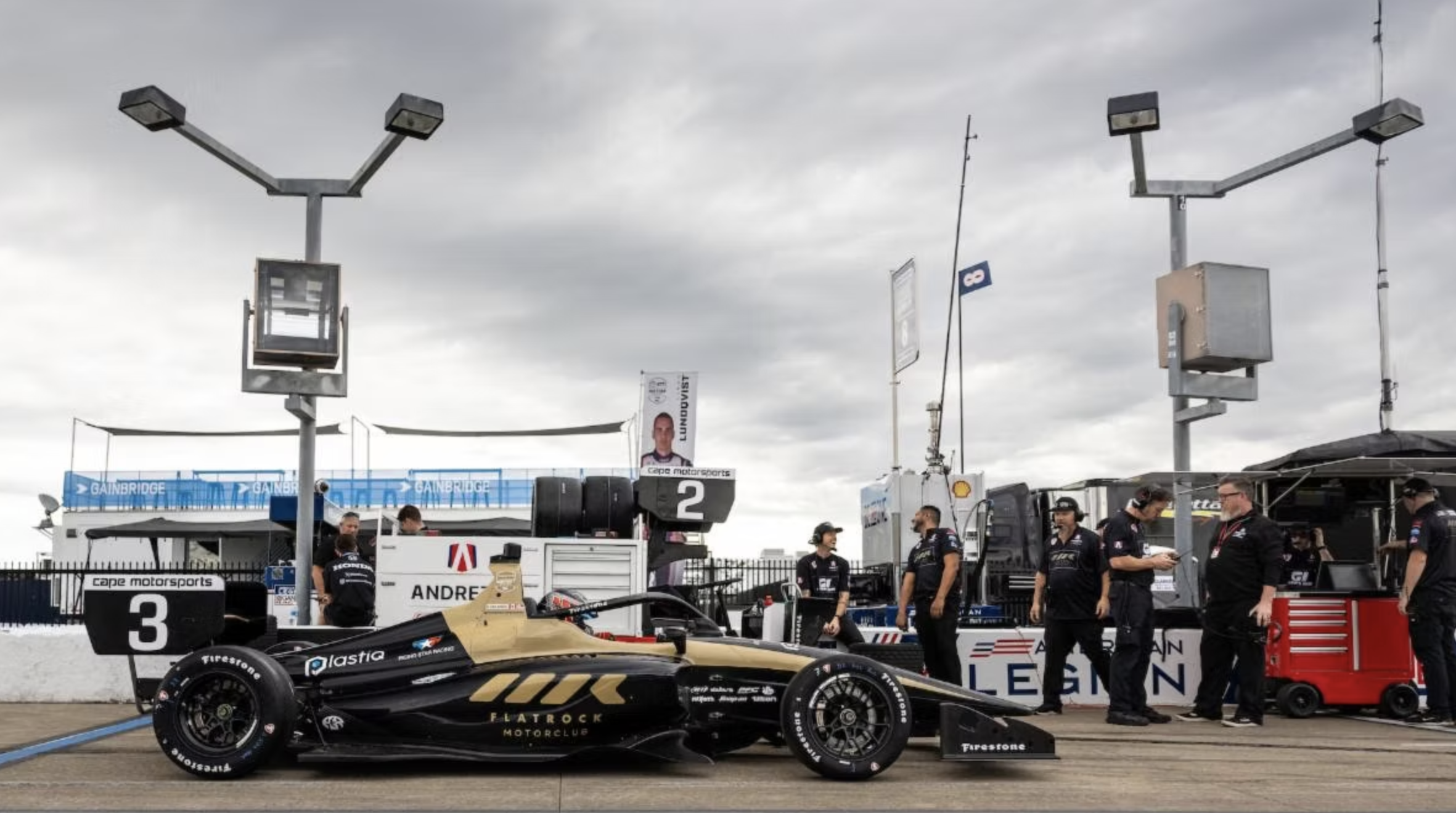 Michael d'Orlando in a pit lane with team members working, race officials, and equipment under cloudy sky at a motorsport event.