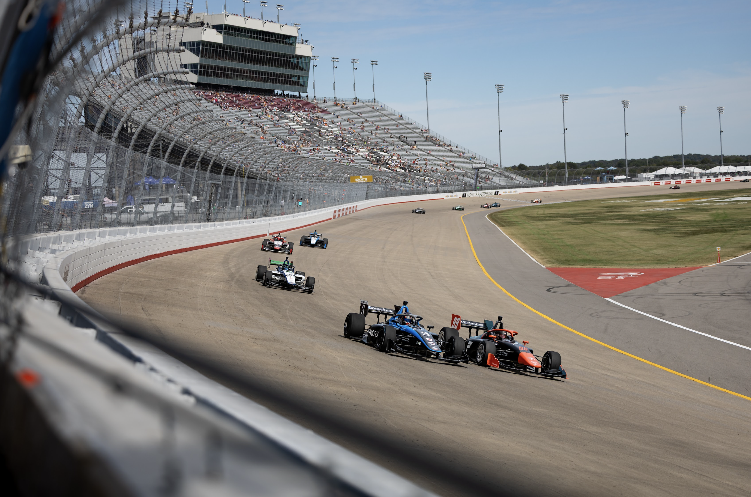 Michael d'Orlando race cars competing on a speedway track with spectators in the grandstands and a large control tower in the background.