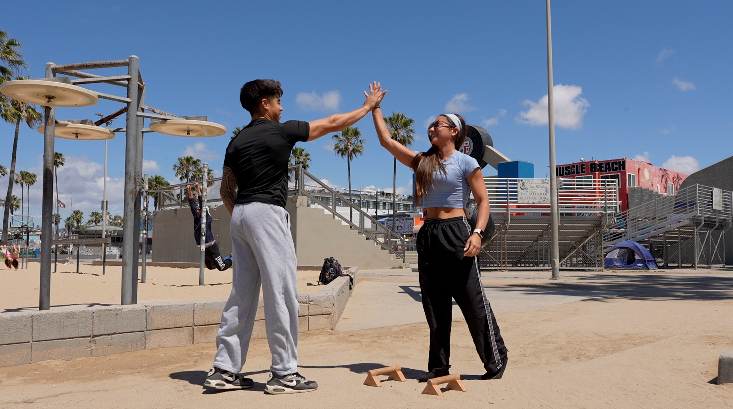 Two people are high-fiving at a beachside workout area with workout equipment, palm trees, and a building labeled 'Muscle Beach' in the background, under a blue sky with a few clouds.