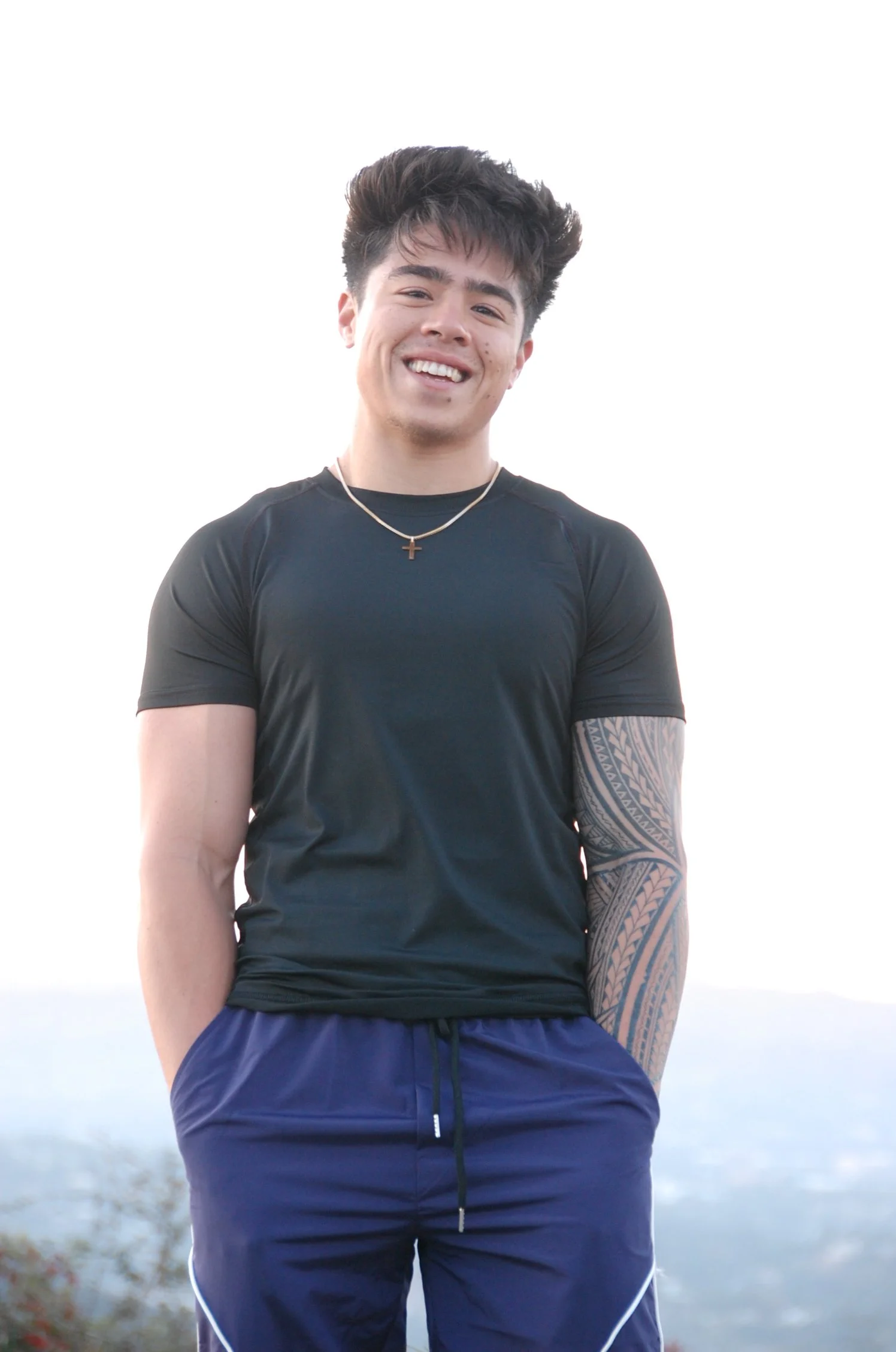 A young man smiling, wearing a black athletic shirt, blue athletic shorts, a gold cross necklace, and having a large tattoo on his left arm, standing outdoors with a blurred natural background.