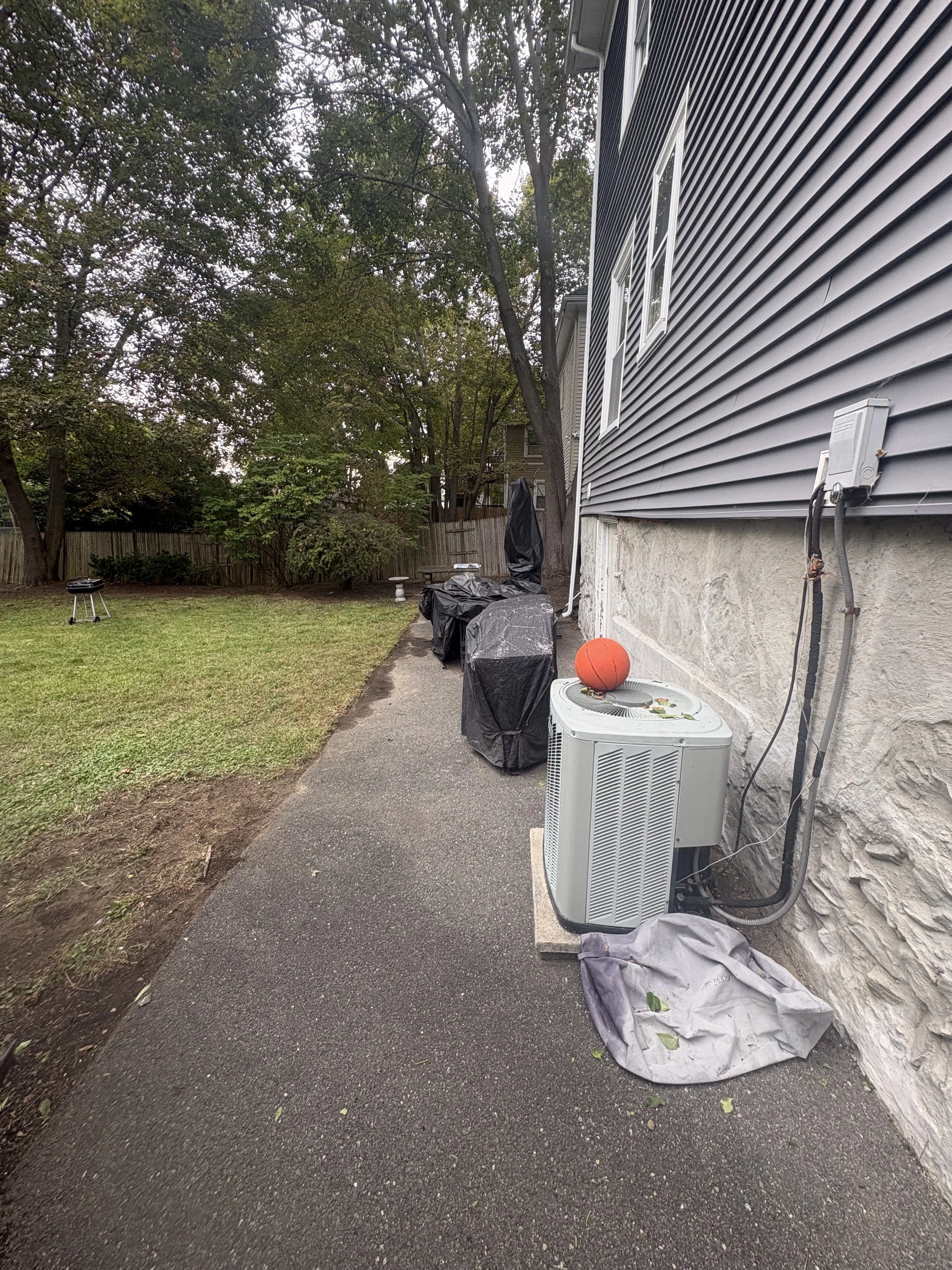 Backyard with trees, wooden fence, patio, covered furniture, air conditioning unit, basketball, and sidewalk.