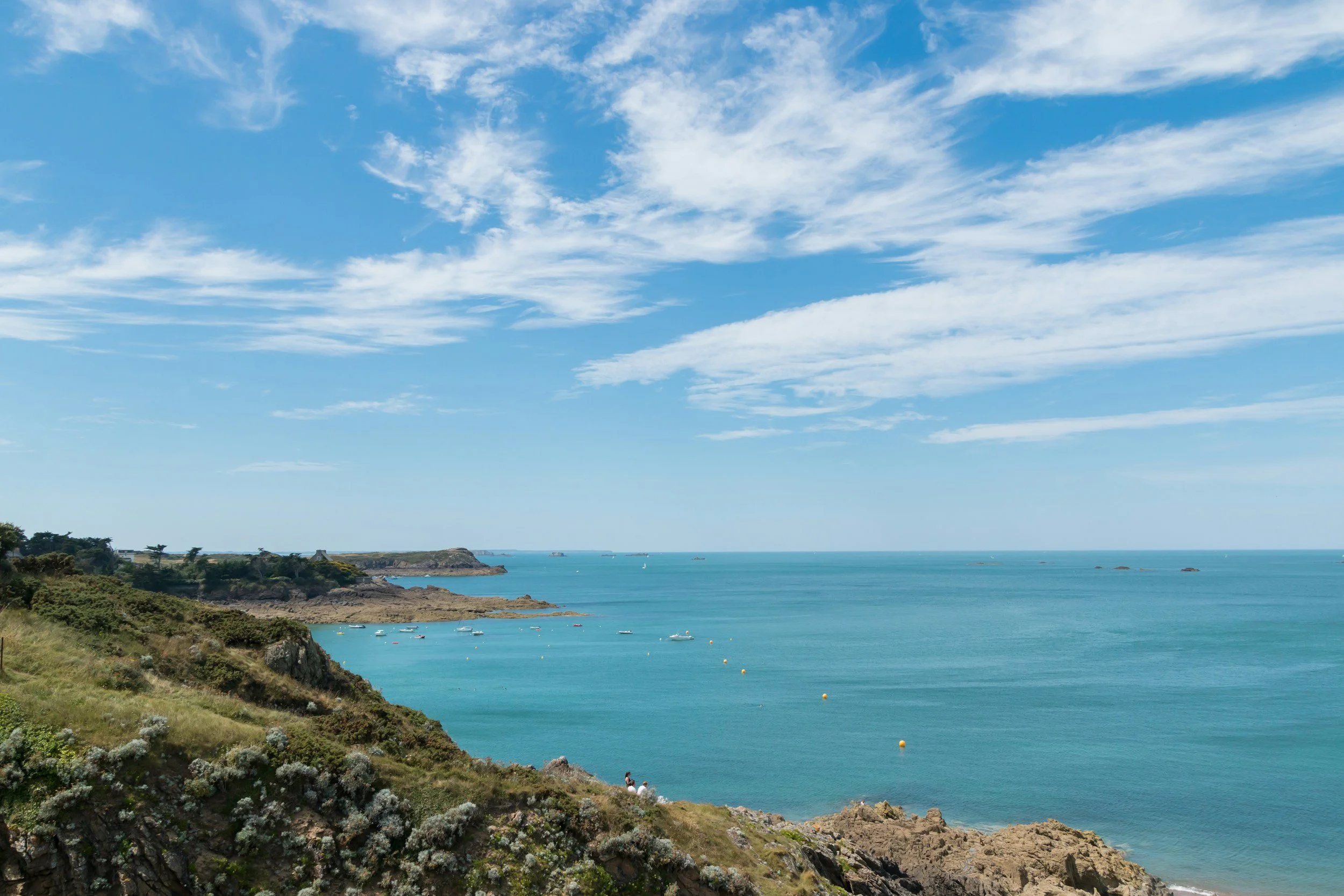 A scenic coastal landscape featuring a bright blue sky with scattered white clouds. In the foreground, lush green vegetation and rocky cliffs overlook a calm, turquoise ocean with several small boats anchored near the shoreline. Distant landforms and trees are visible along the coast.