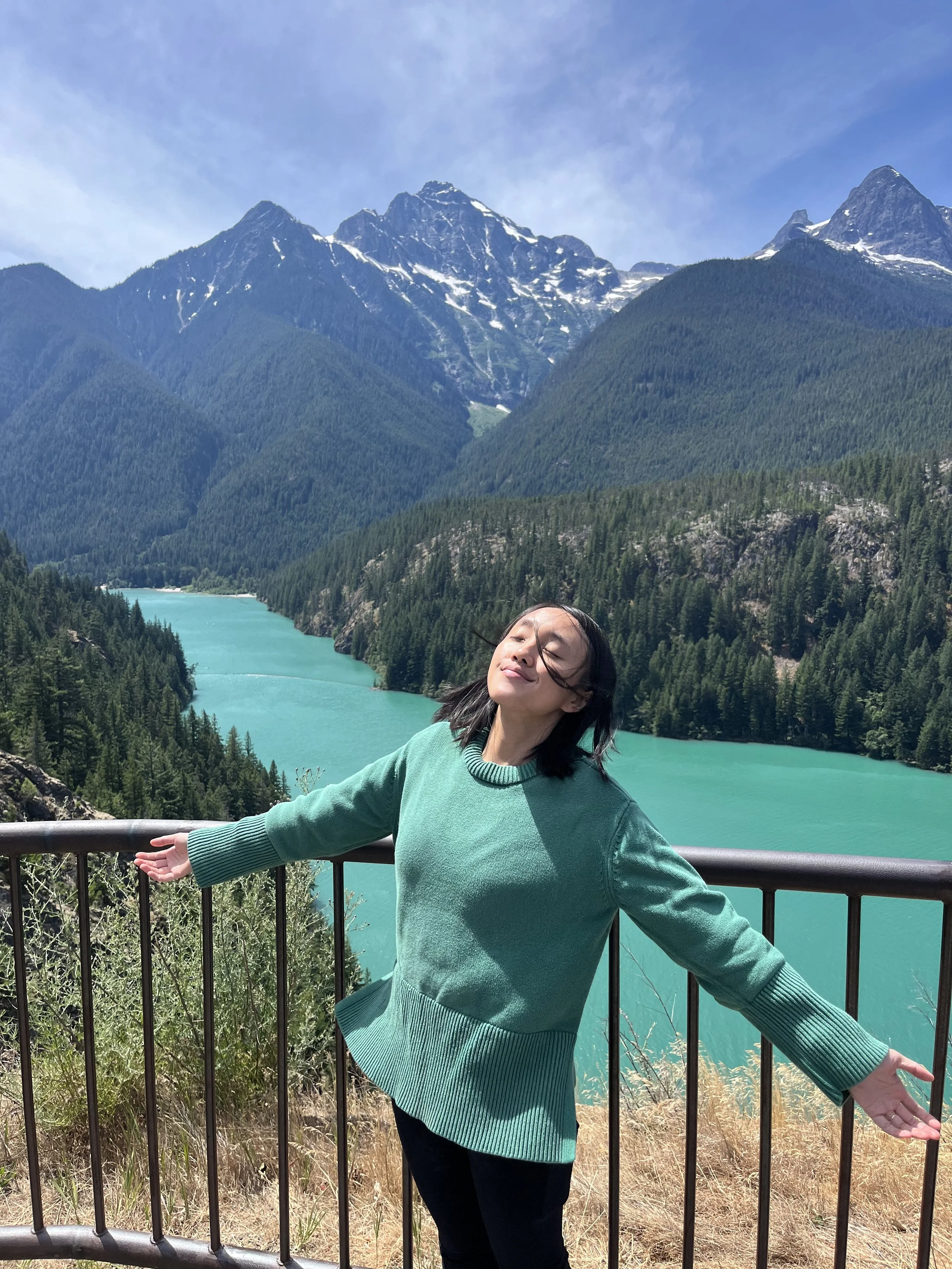 A woman in a green sweater stands with arms outstretched in front of a scenic mountain landscape with a turquoise lake and forested mountains in the background.