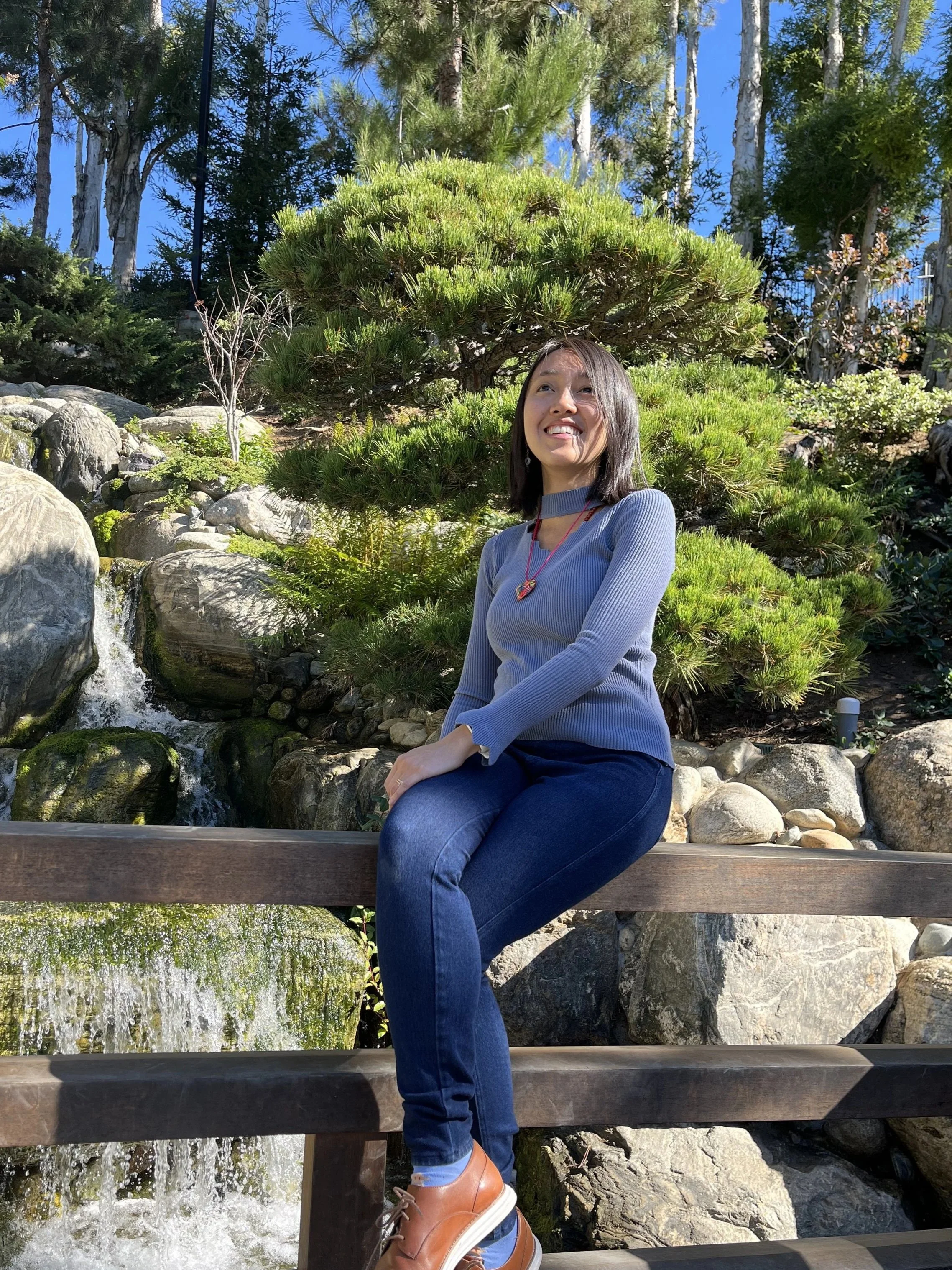 A woman sitting on a wooden bench outdoors in a garden, with green shrubs, rocks, and a small waterfall behind her, smiling and looking up.
