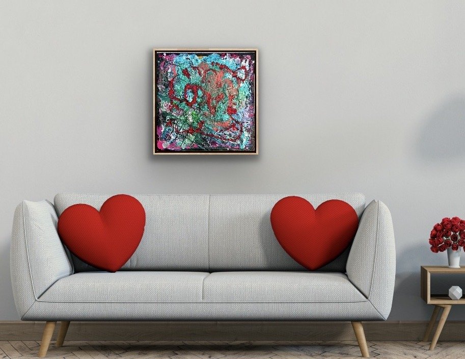 A modern living room with a white sofa decorated with two large red heart-shaped pillows. There is a small side table with a vase of red flowers on the right. An abstract colorful painting hangs on the wall above the sofa.