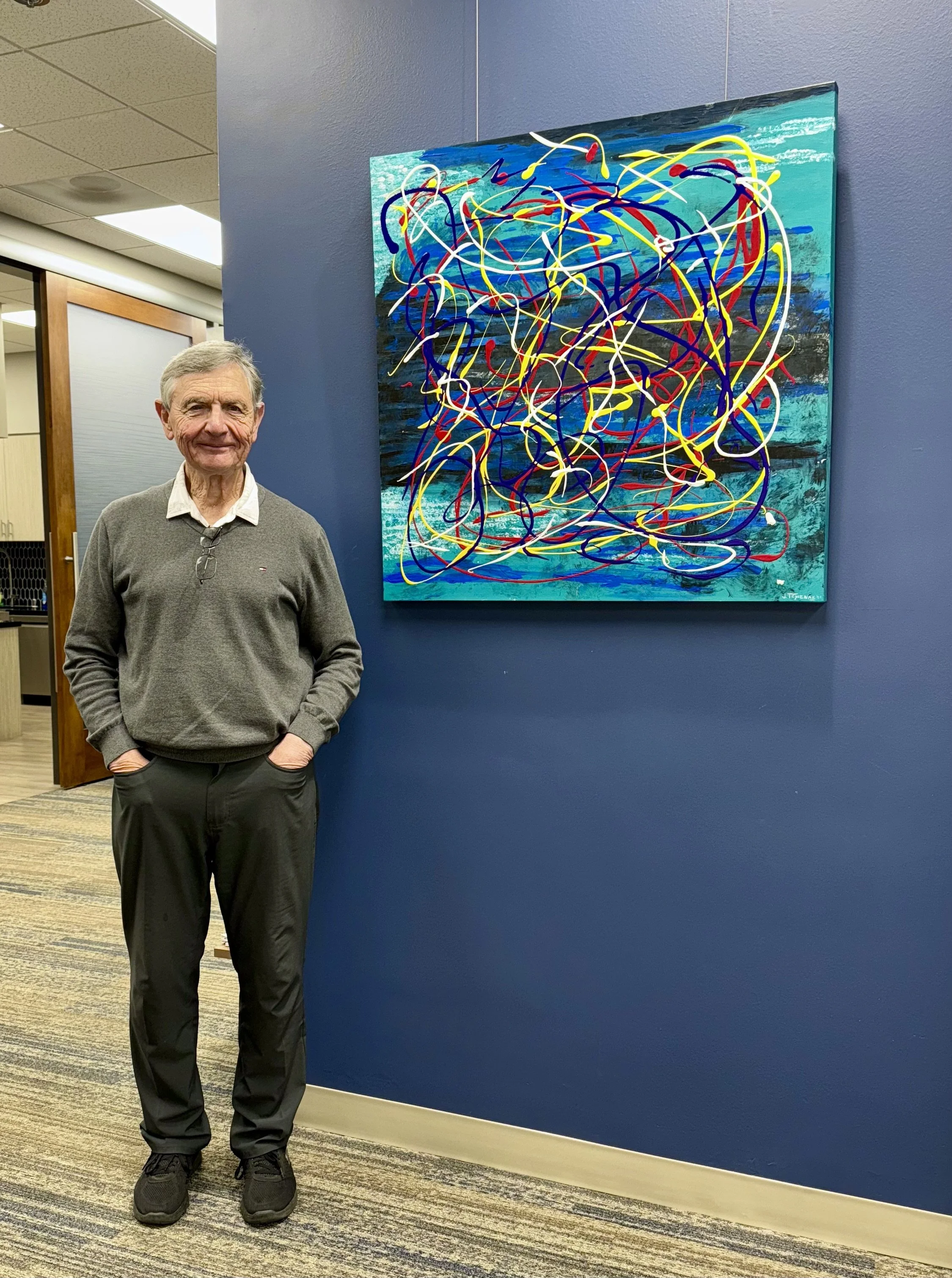 An elderly man wearing a grey sweater, black pants, and black athletic shoes standing in front of a colorful abstract painting on a deep blue wall.