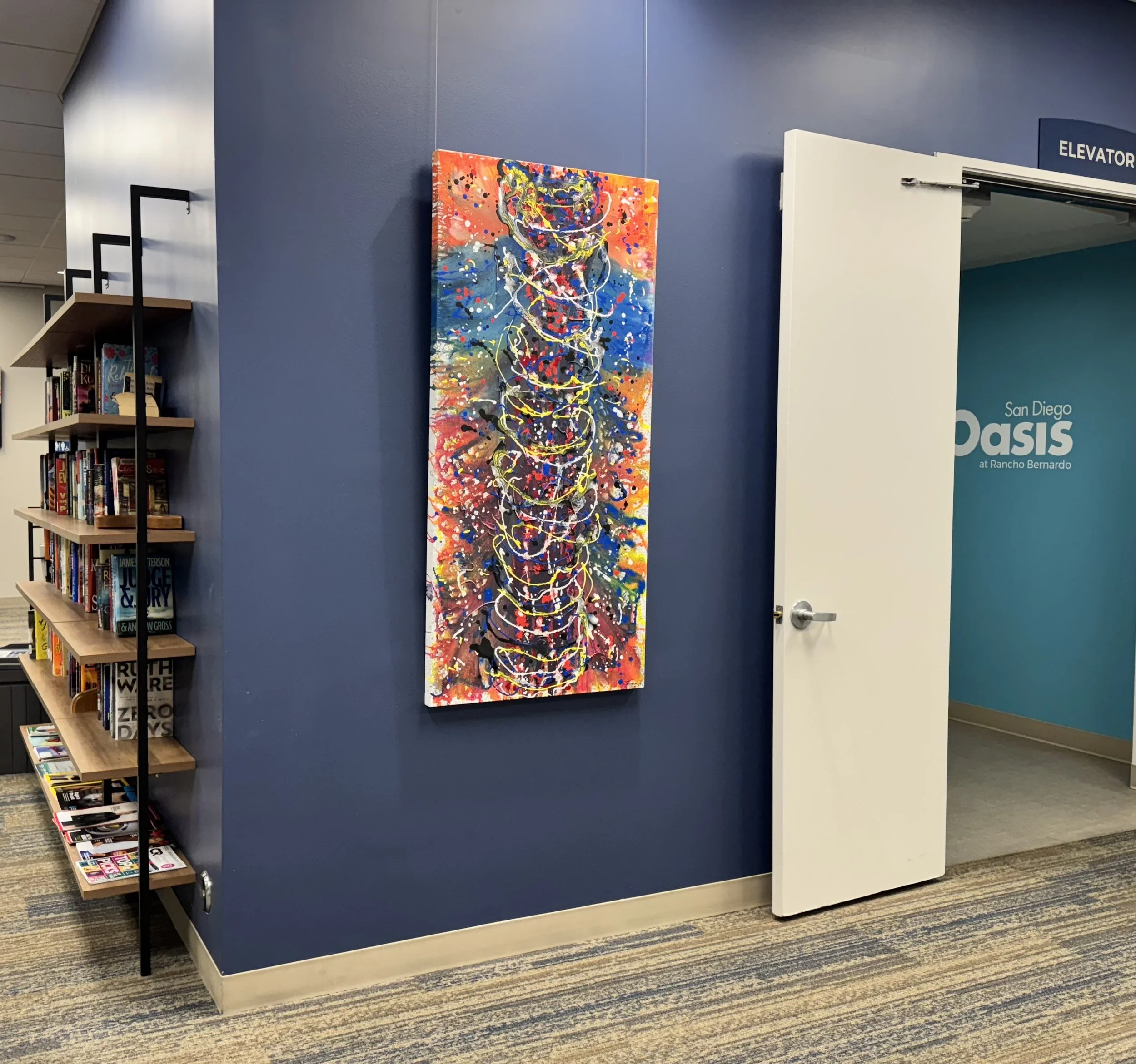 Colorful abstract painting on a blue wall in a library or bookstore, with a bookshelf filled with books on the side and a white door with a sign that says 'ELEVATOR' and another sign indicating it is at Rancho Bernardo, San Diego.
