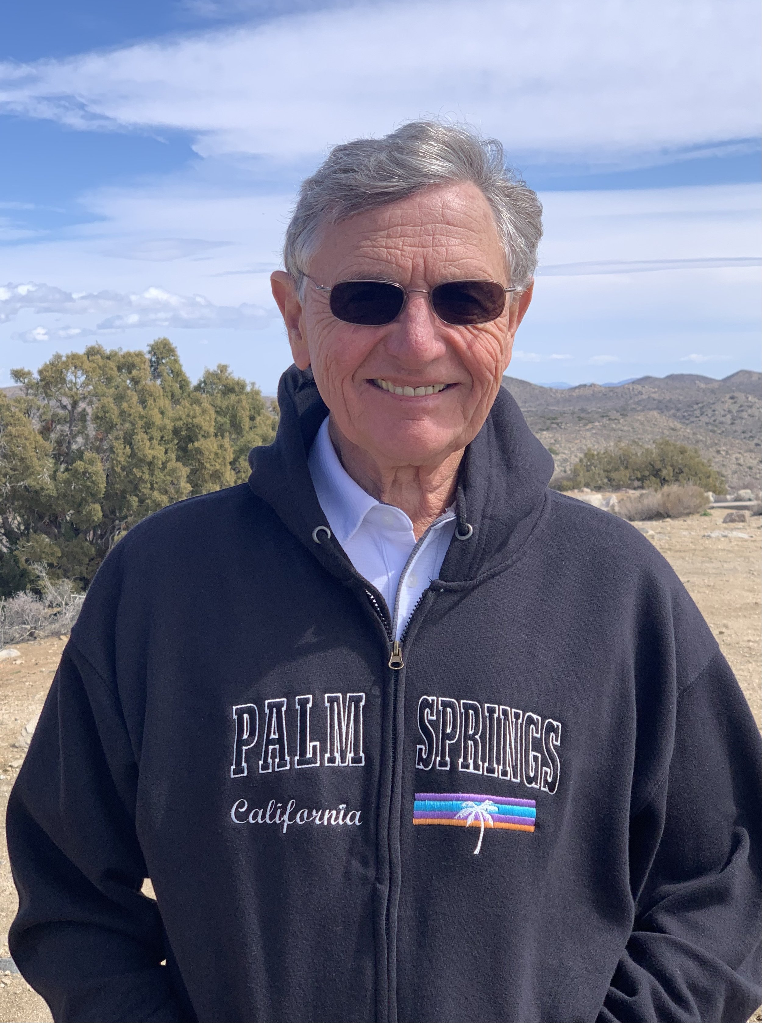 Man with gray hair, wearing sunglasses, smiling, dressed in a black Palm Springs California jacket with a sunset and palm tree graphic, standing outdoors with mountains, trees, and a partly cloudy sky in the background.