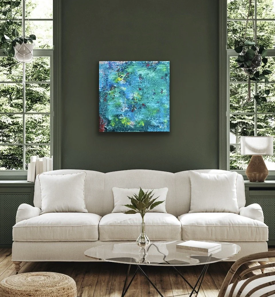 A living room with a white sofa, a glass coffee table with a plant, a bookshelf, a lamp, and large windows showing green trees outside. There is a colorful abstract painting on the dark green wall behind the sofa.
