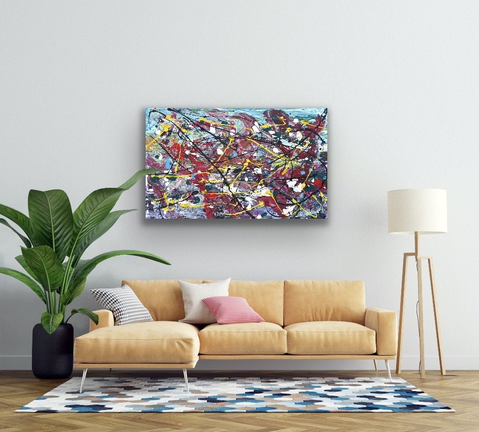 Living room with a light brown sofa, colorful cushions, an abstract painting on the white wall, a large potted plant to the left, a tall floor lamp to the right, and a geometric-patterned rug on the wooden floor.