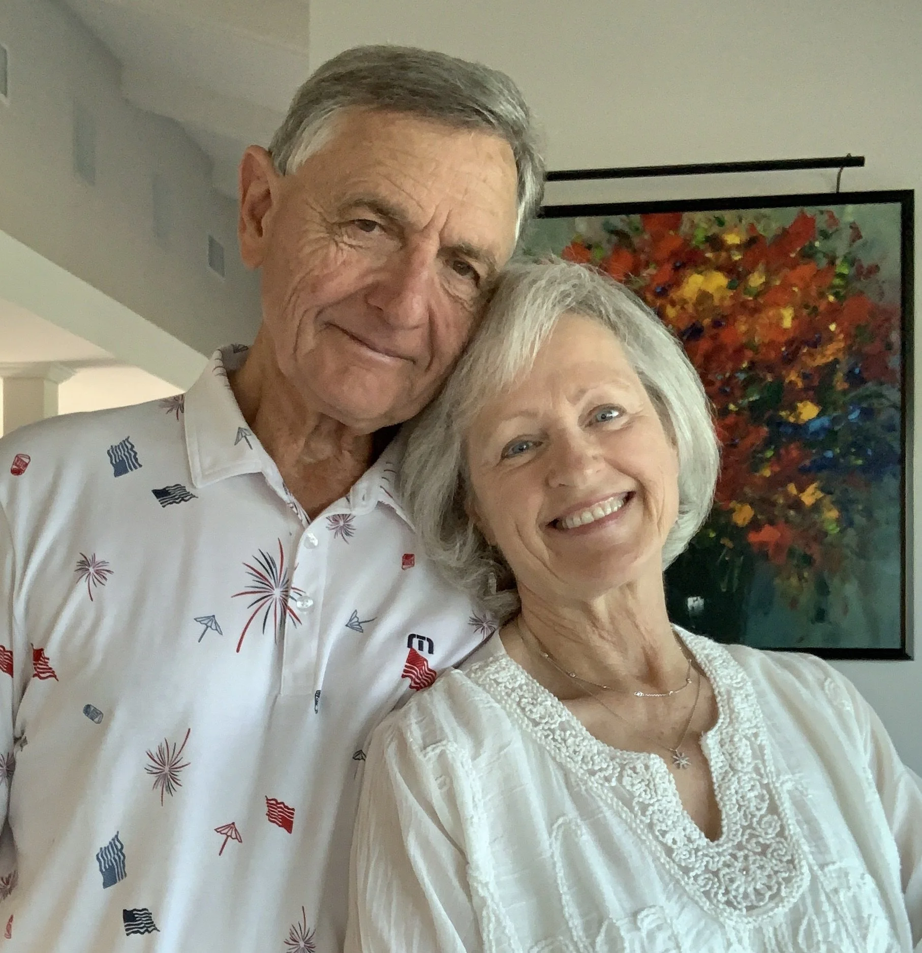 An elderly couple smiling and hugging indoors, with a colorful abstract painting in the background.
