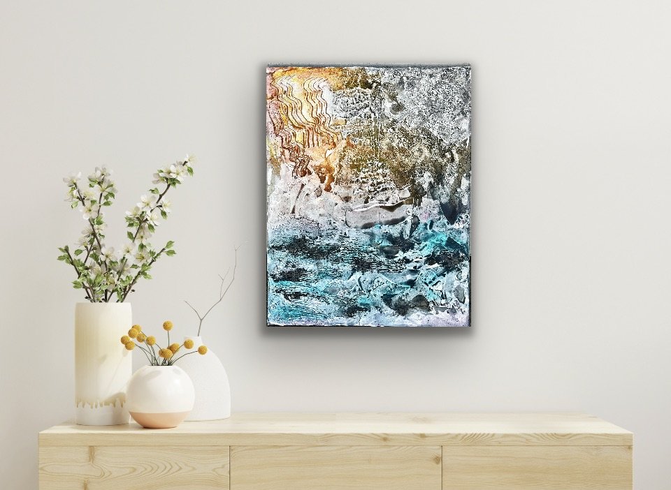 Abstract painting with textured patterns in shades of gold, gray, black, and blue hanging on a plain white wall.
