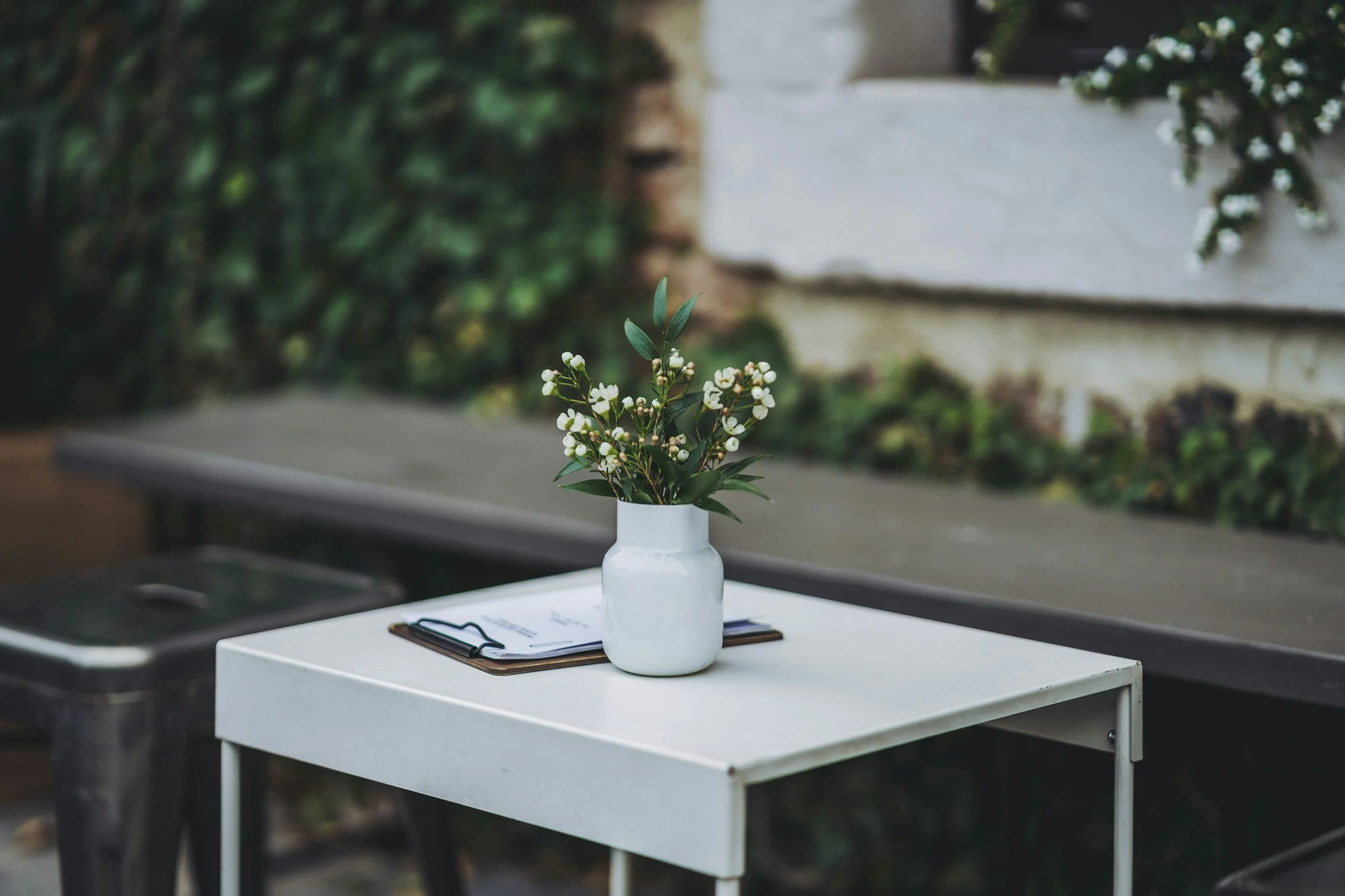 A white table with a white vase holding green leafy stems and small white flowers, a closed black notebook with a clipboard on top, and a pen on the clipboard, with a background of green trees and a white wall.