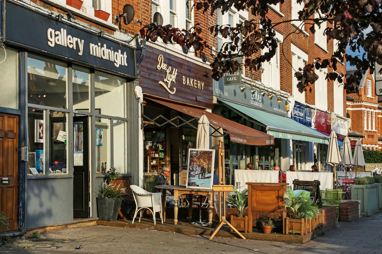 Street view of a row of cafes and shops with outdoor seating, including 'Gallery Midnight', 'Dee Light Bakery', and other establishments, with people sitting outside, plants, and umbrellas on a sunny day.