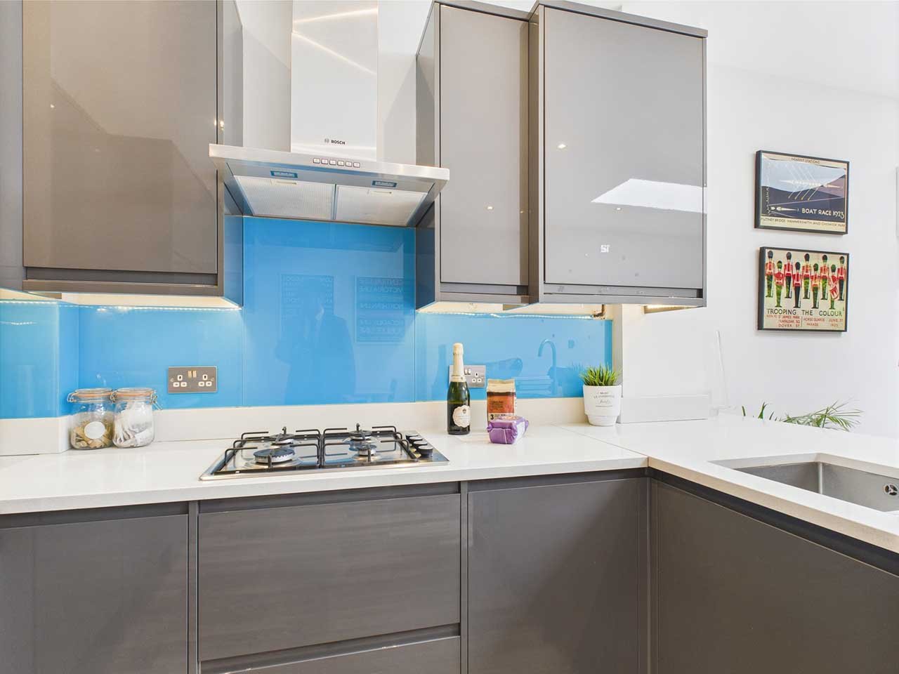 Modern kitchen with gray cabinets, white countertop, blue glass backsplash, gas stove, wall-mounted range hood, small potted plant, bottles, and framed artwork on the wall.