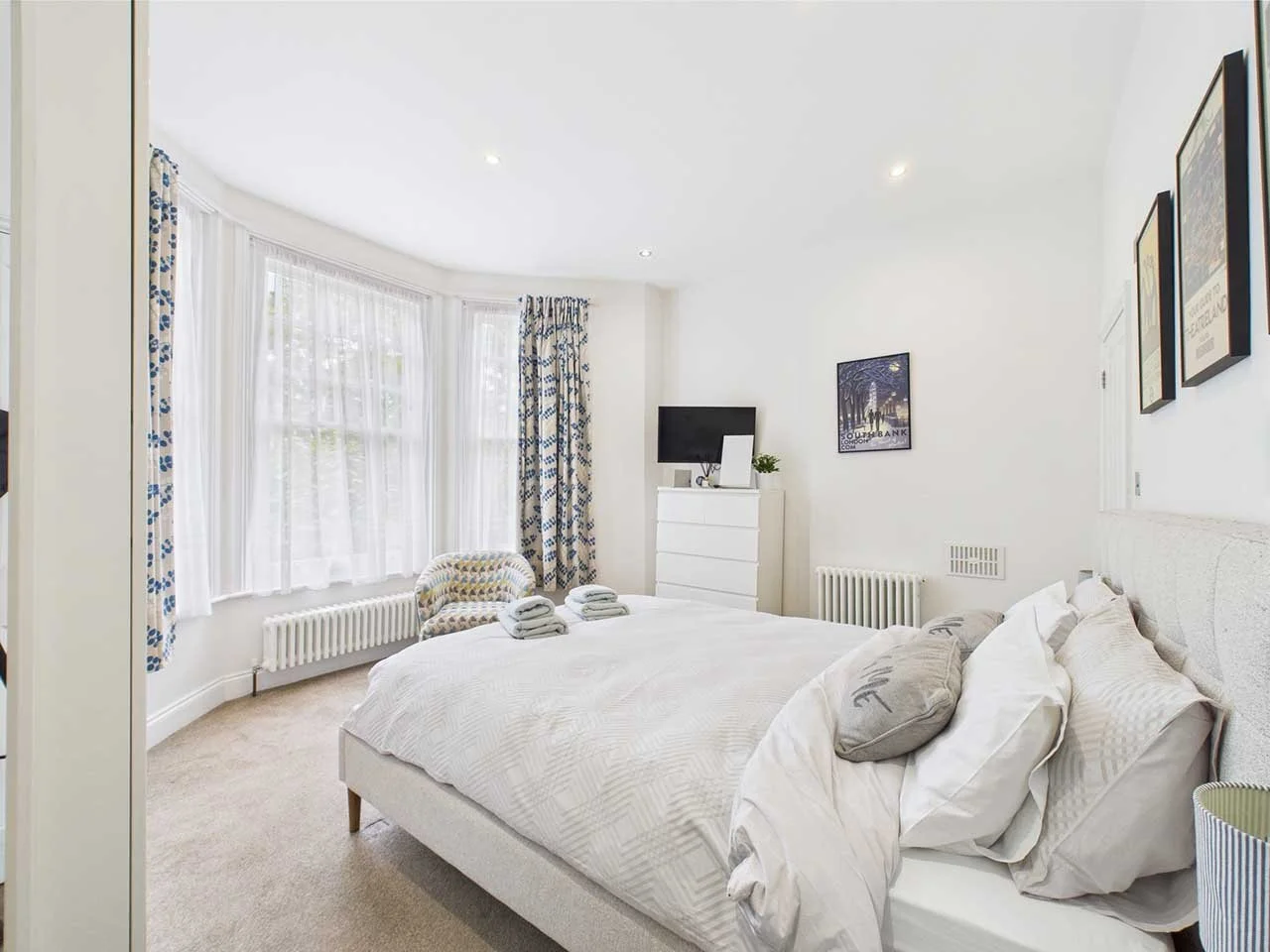 Bright bedroom with white walls, large bay windows with sheer and patterned curtains, neatly made bed with multiple pillows, and a dresser with a small television and a potted plant.