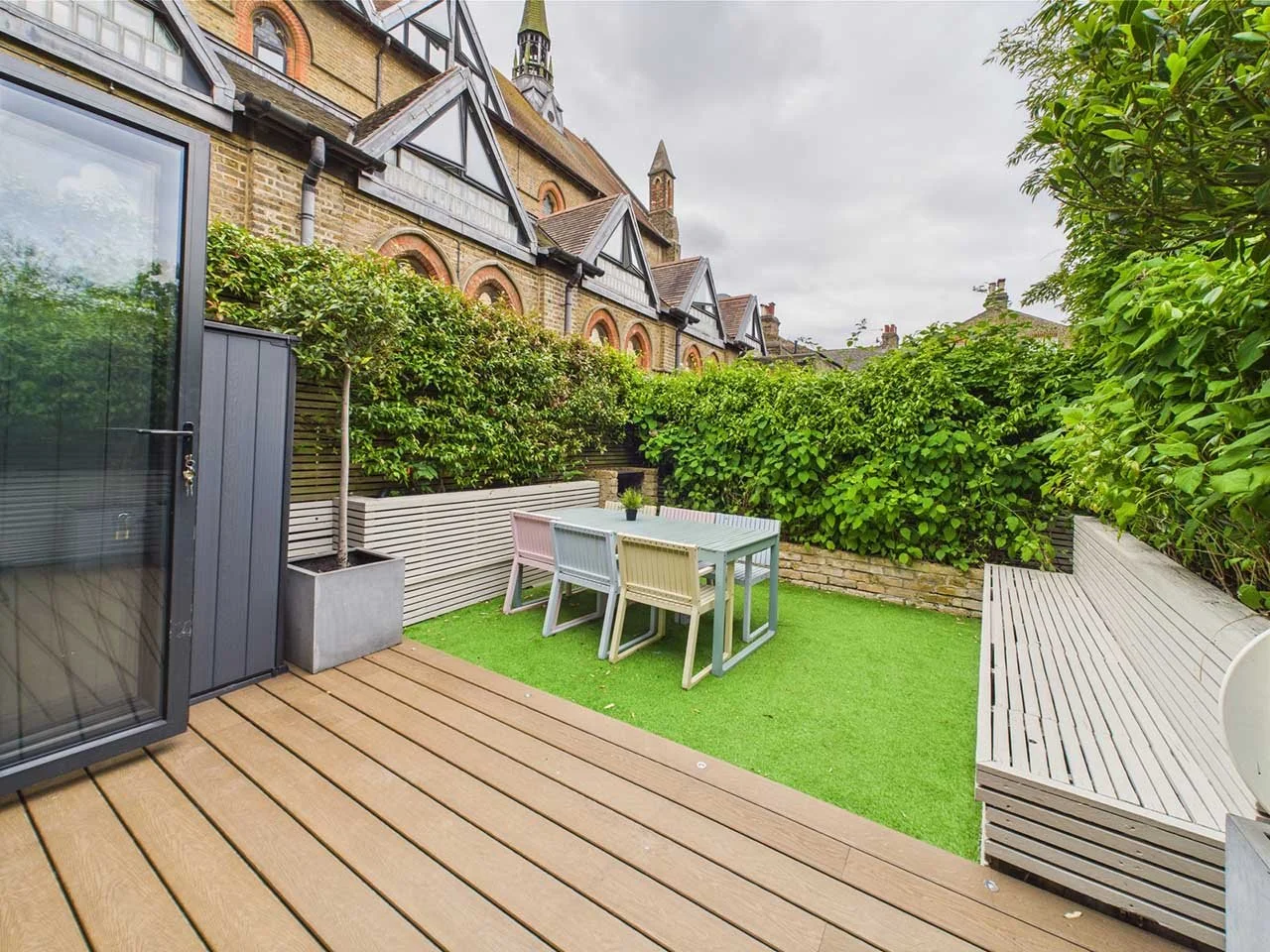 Outdoor backyard patio with wooden decking, a small table with four chairs, a lush green hedge, and a bench, surrounded by a brick wall and neighboring house buildings, under an overcast sky.