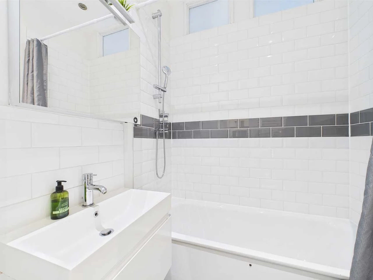 A modern bathroom with white subway tiles and a gray tile accent strip, featuring a bathtub, a sink with a green soap bottle, a mirror, a showerhead, and a window.
