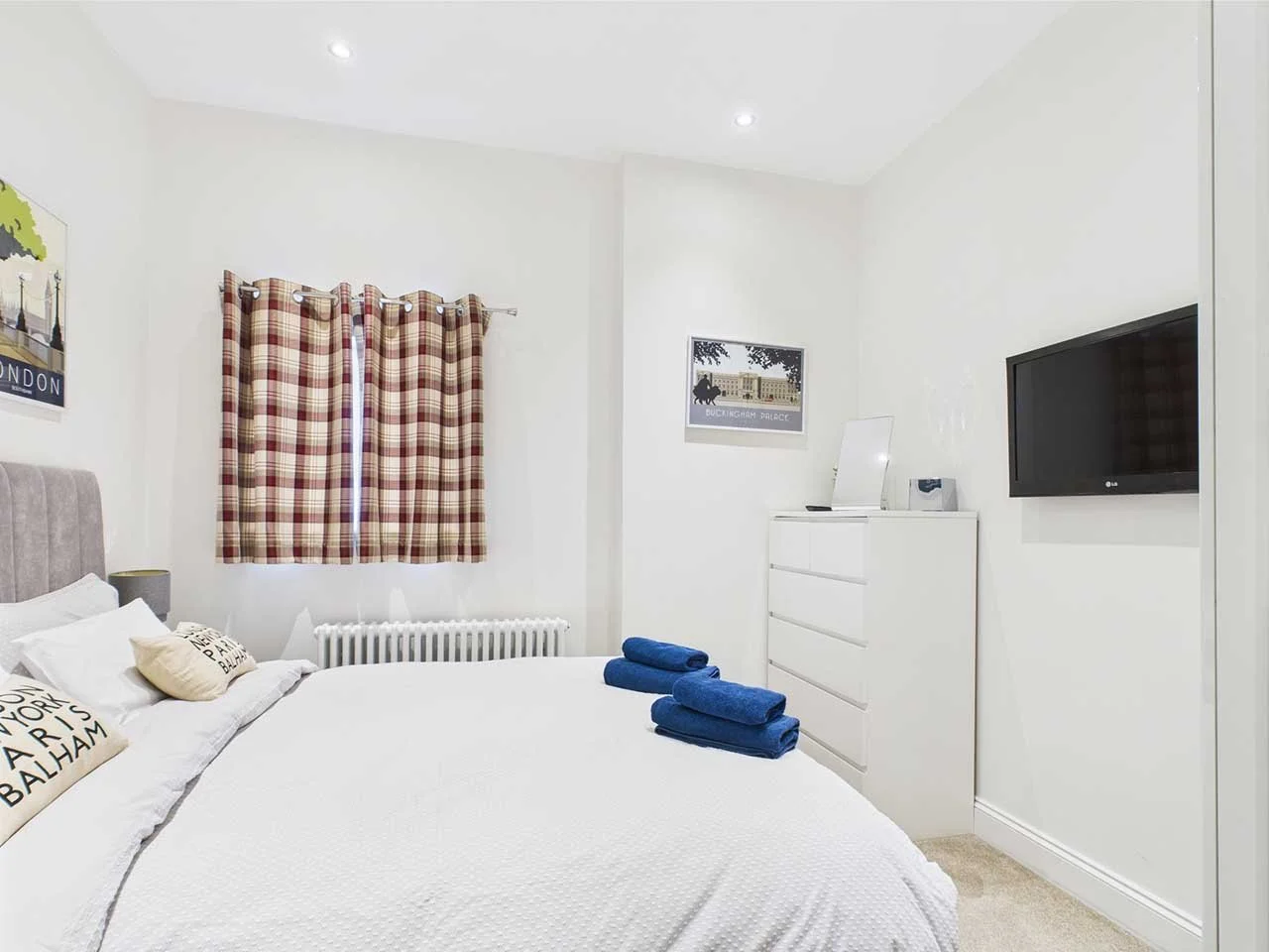 A clean, white bedroom with a bed, curtains, artwork, a dresser, and a wall-mounted television.
