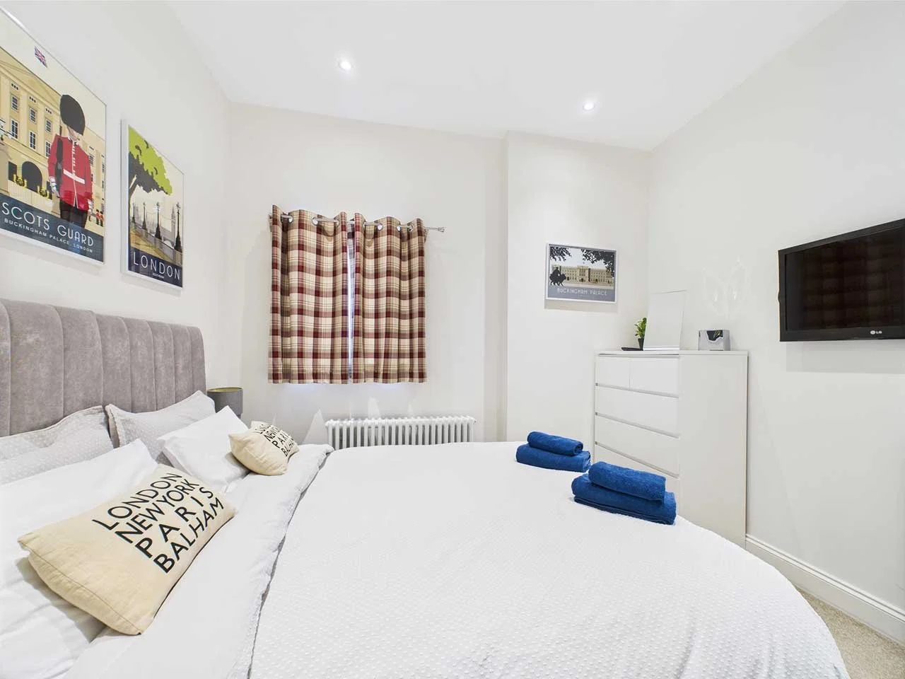 A bright, minimalist bedroom with a bed, checkered curtains, wall art, a white dresser, and a flat-screen TV.