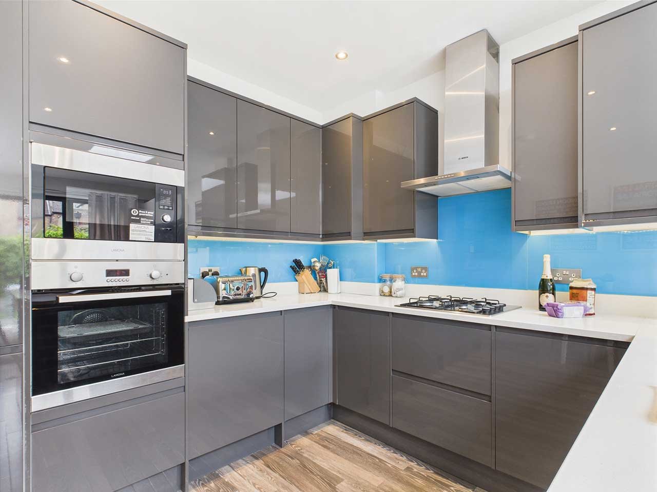 Modern kitchen with gray cabinets, white countertops, blue backsplash, built-in oven and microwave, stove, stainless steel range hood, and various kitchen accessories.