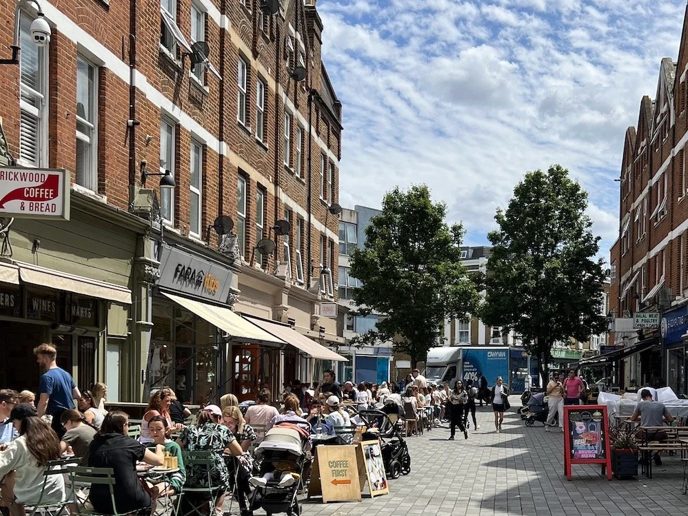 Outdoor dining on a busy city street with many people sitting at tables, surrounded by brick buildings, trees, and a blue sky with clouds.