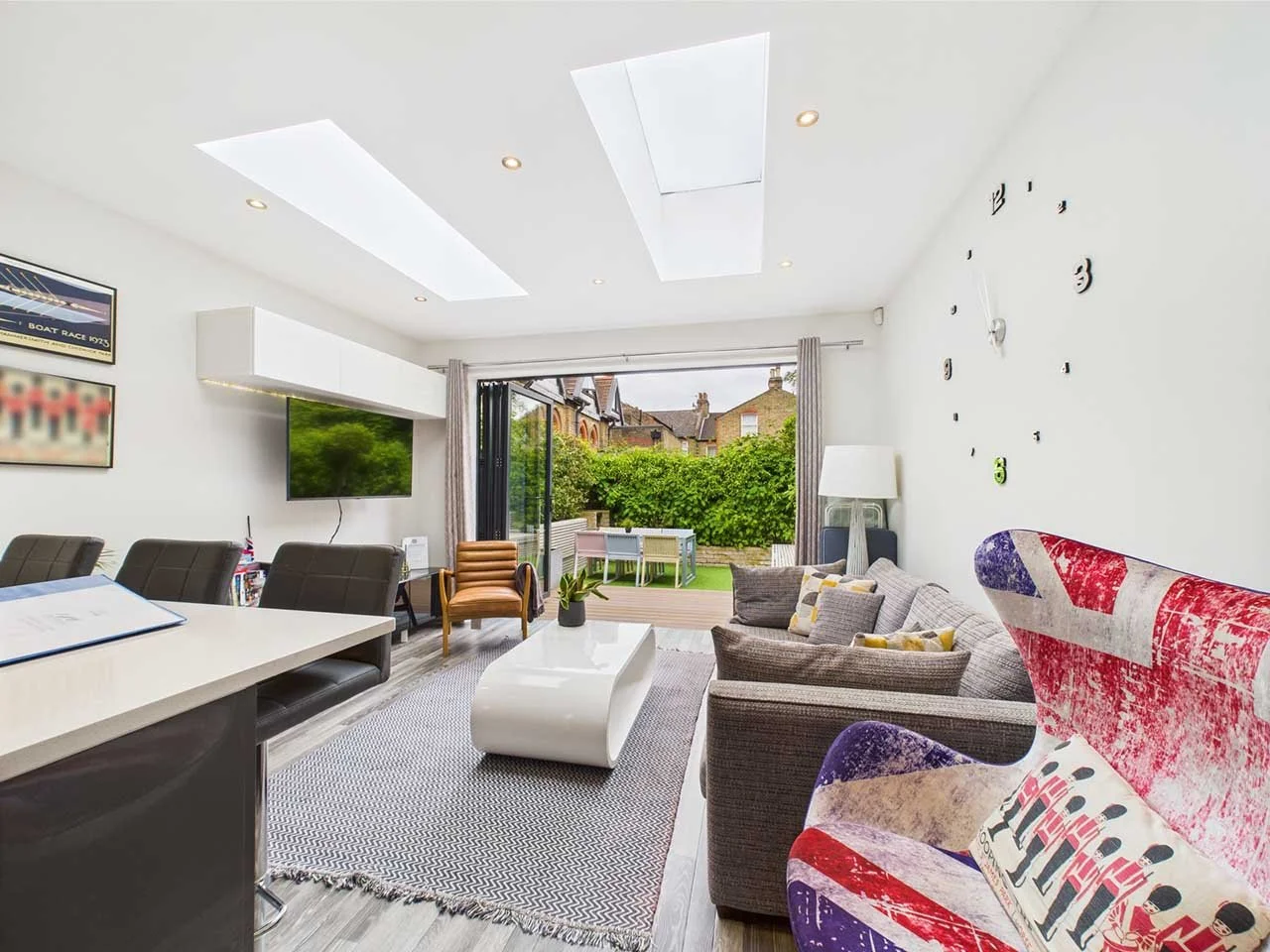 Bright living room with gray sofas, a white coffee table, and a large wall clock, opening to a patio with outdoor seating and a garden.