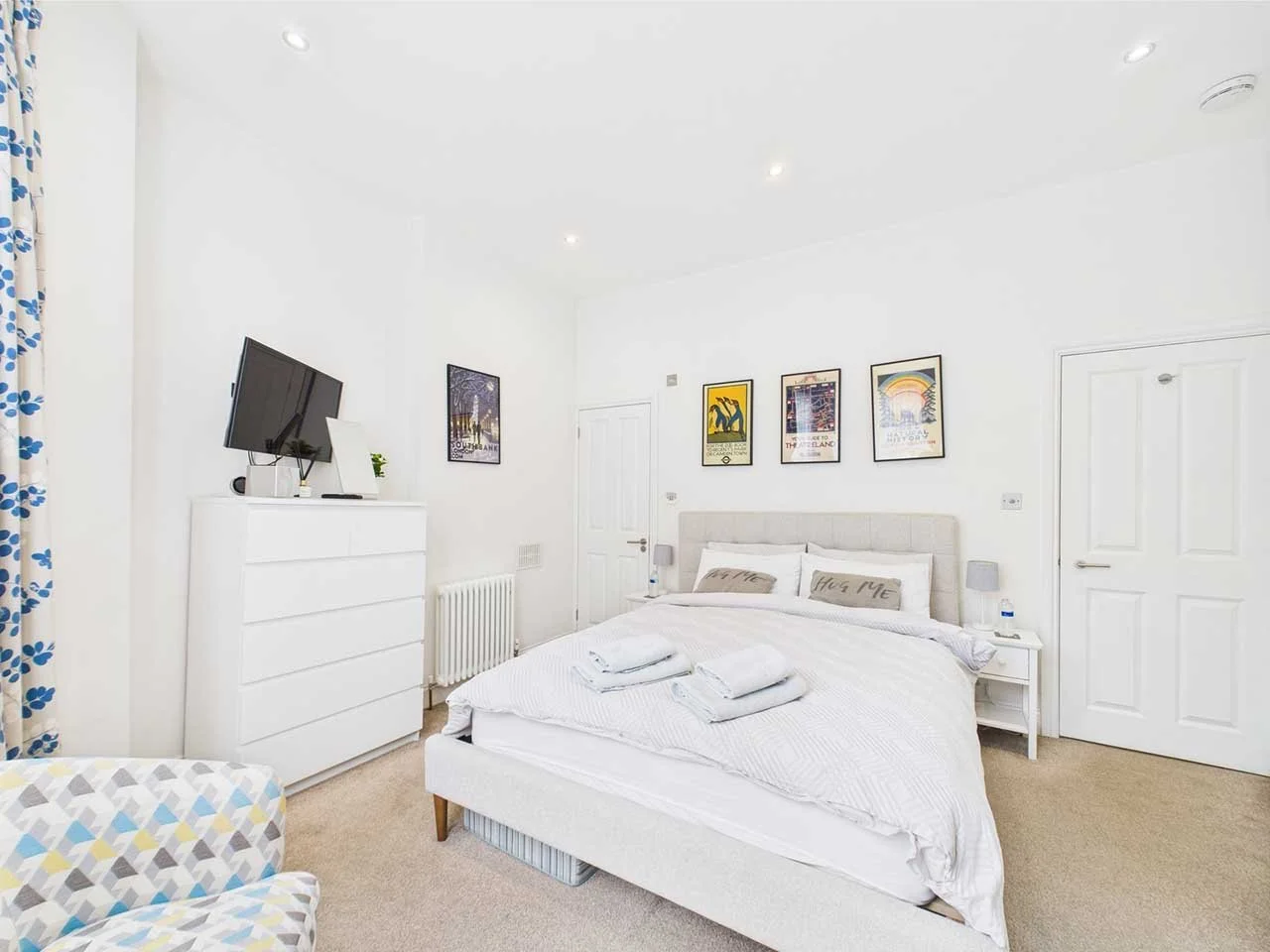 A bright modern bedroom with a white bed, bedside tables, and framed artwork on the wall. Includes a dresser with a TV and a patterned armchair.