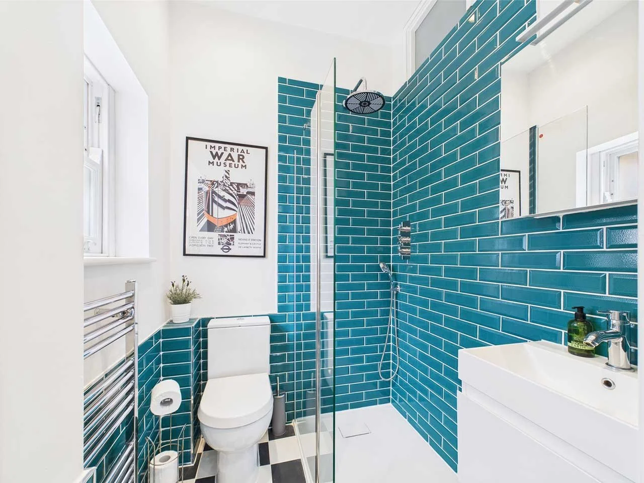 Bathroom with white walls and a blue tiled shower area. There is a toilet, a showerhead, a mirror, a small sink with a soap dispenser, a framed picture on the wall, a window, and a towel rack.