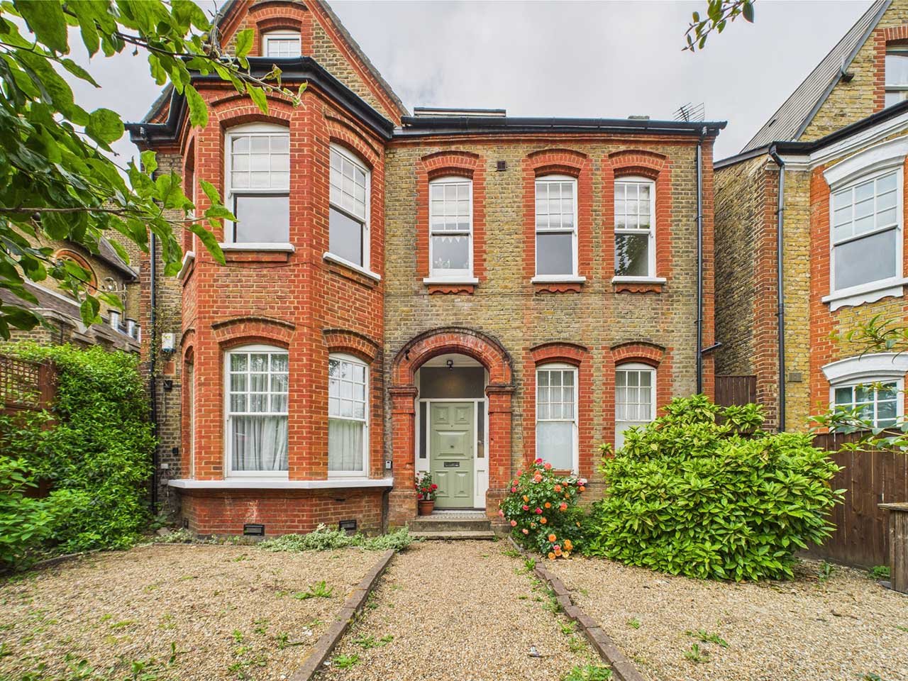 Three-story brick residential house with arched windows and a green front door, surrounded by a small front garden with bushes and flowers.