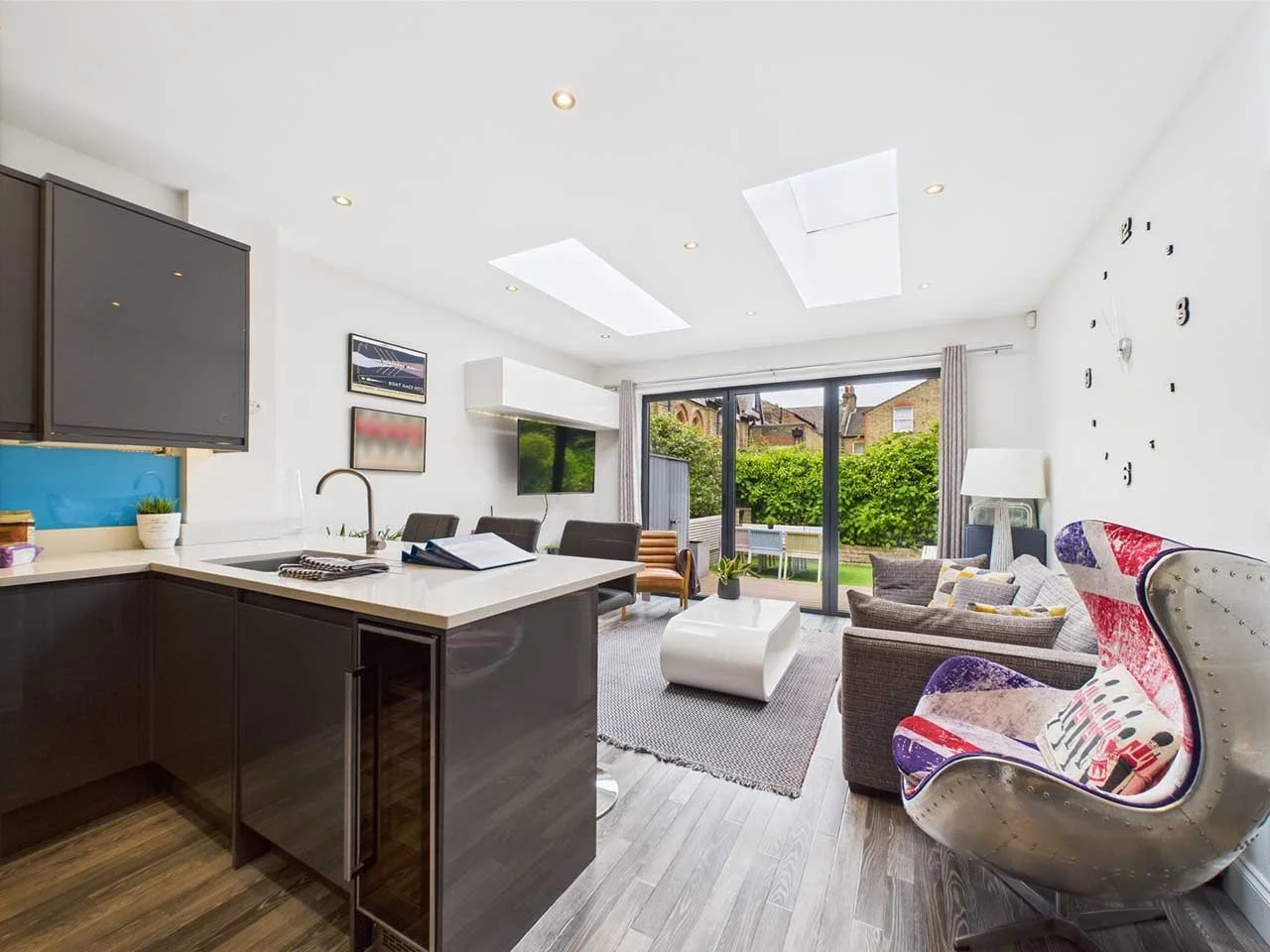 Modern open-concept living room and kitchen with gray and white color scheme, large sliding glass doors leading to a backyard garden, stylish furniture, and contemporary decor.