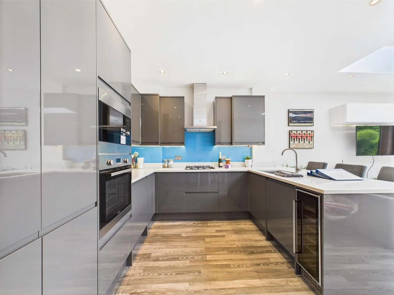 Modern kitchen with grey cabinets, white countertops, blue backsplash, and wooden flooring. Includes built-in oven, gas stove, and glass-front wine cooler.
