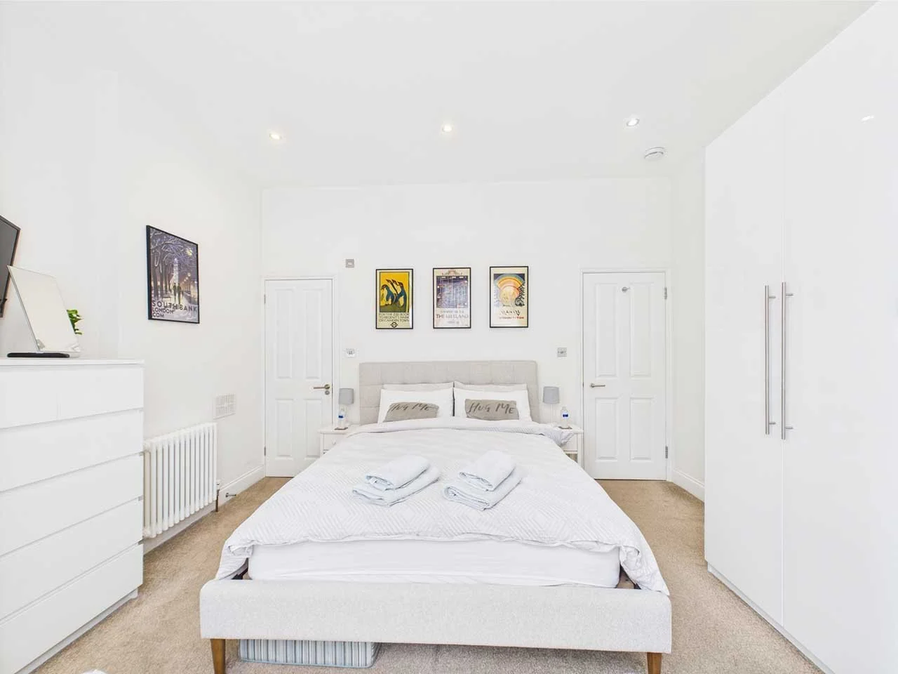 A bright white bedroom with a bed centered against the wall, decorated with white linens and pillows. There are three framed posters above the bed and towels laid out on the bed. White furniture, including dressers and a wardrobe, are on each side of