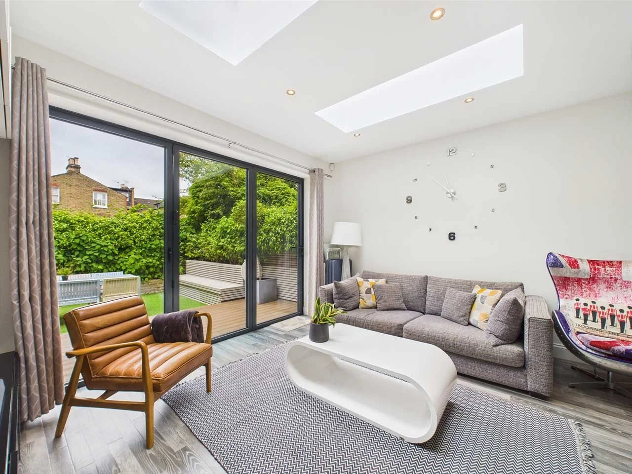 Living room with large sliding glass doors opening to a backyard garden. Contains a gray sectional sofa with cushions, a white modern coffee table, a wooden armchair with a leather seat, a floor lamp, a round wall clock, and a colorful chair with a g