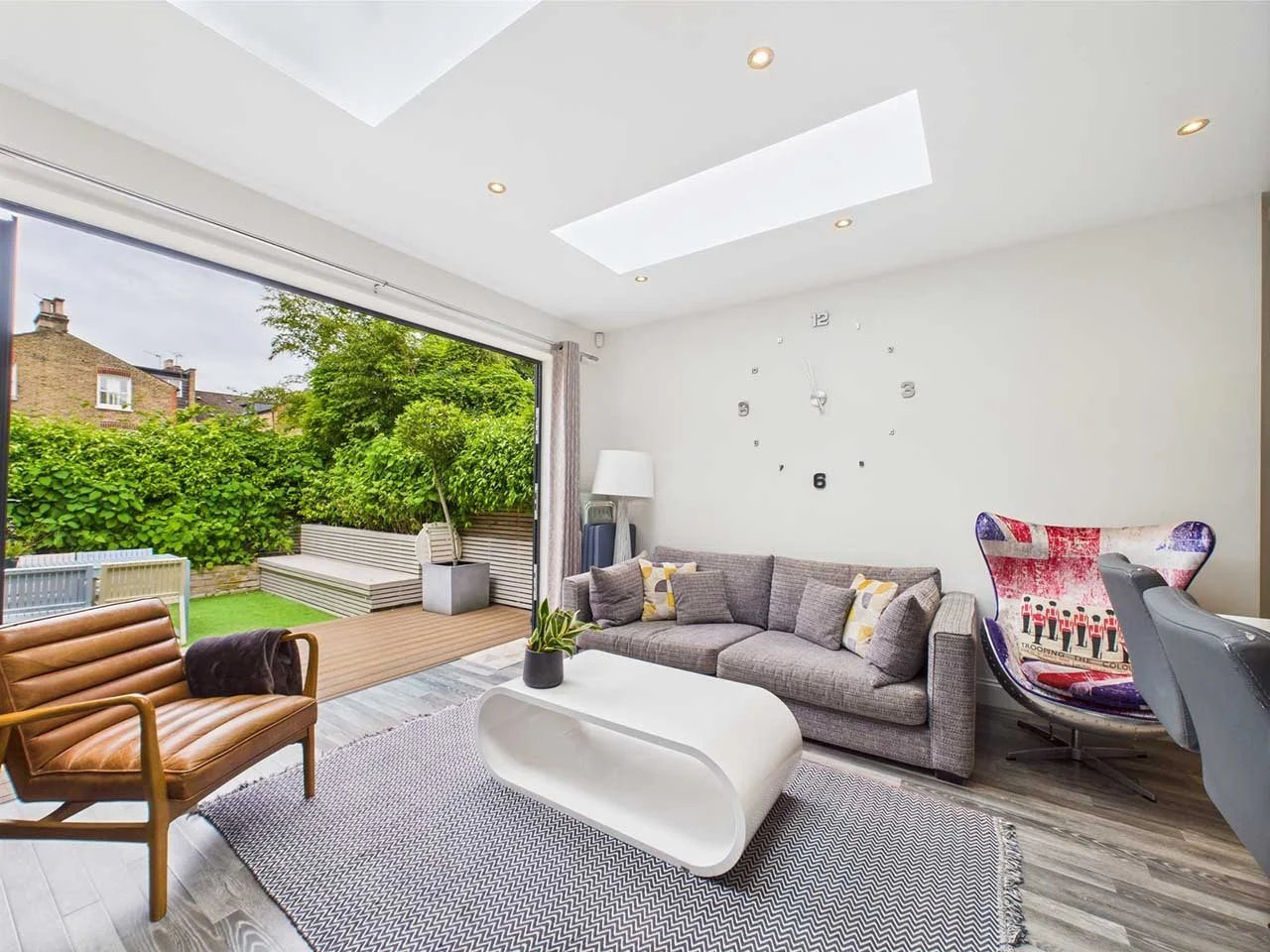 Living room with gray sofa, modern white coffee table, leather armchair, colorful bean bag chair, large open sliding door leading to a backyard with greenery, wooden deck, and outdoor seating, wall clock, ceiling skylights, and wooden flooring.