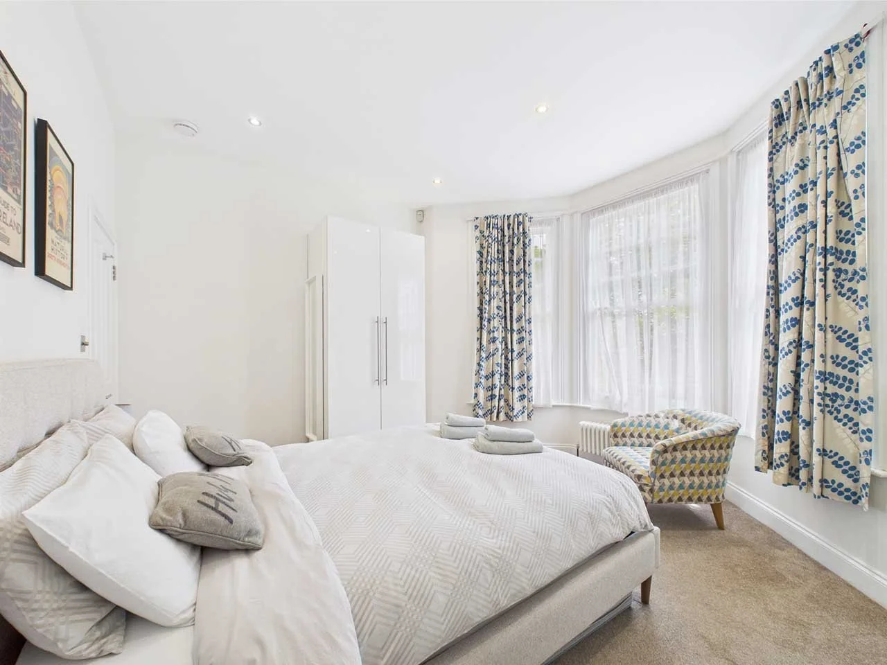 Bright bedroom with a bed, pillows, curtains, a patterned armchair, and artwork on the walls.