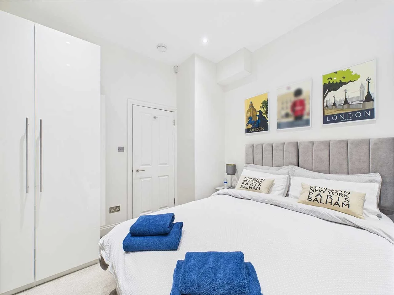Modern white bedroom with a bed, pillows labeled with city names, and artwork featuring London scenes above the headboard