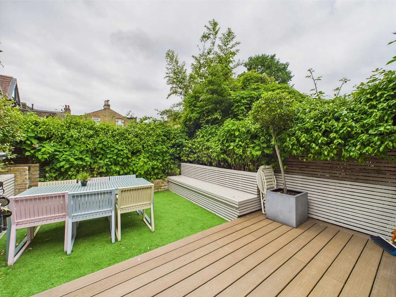 A backyard patio with wooden decking and artificial grass, featuring a pastel-colored dining table with white, pink, and yellow chairs, a potted tree, a white bench, and green bushes along the fence under an overcast sky.