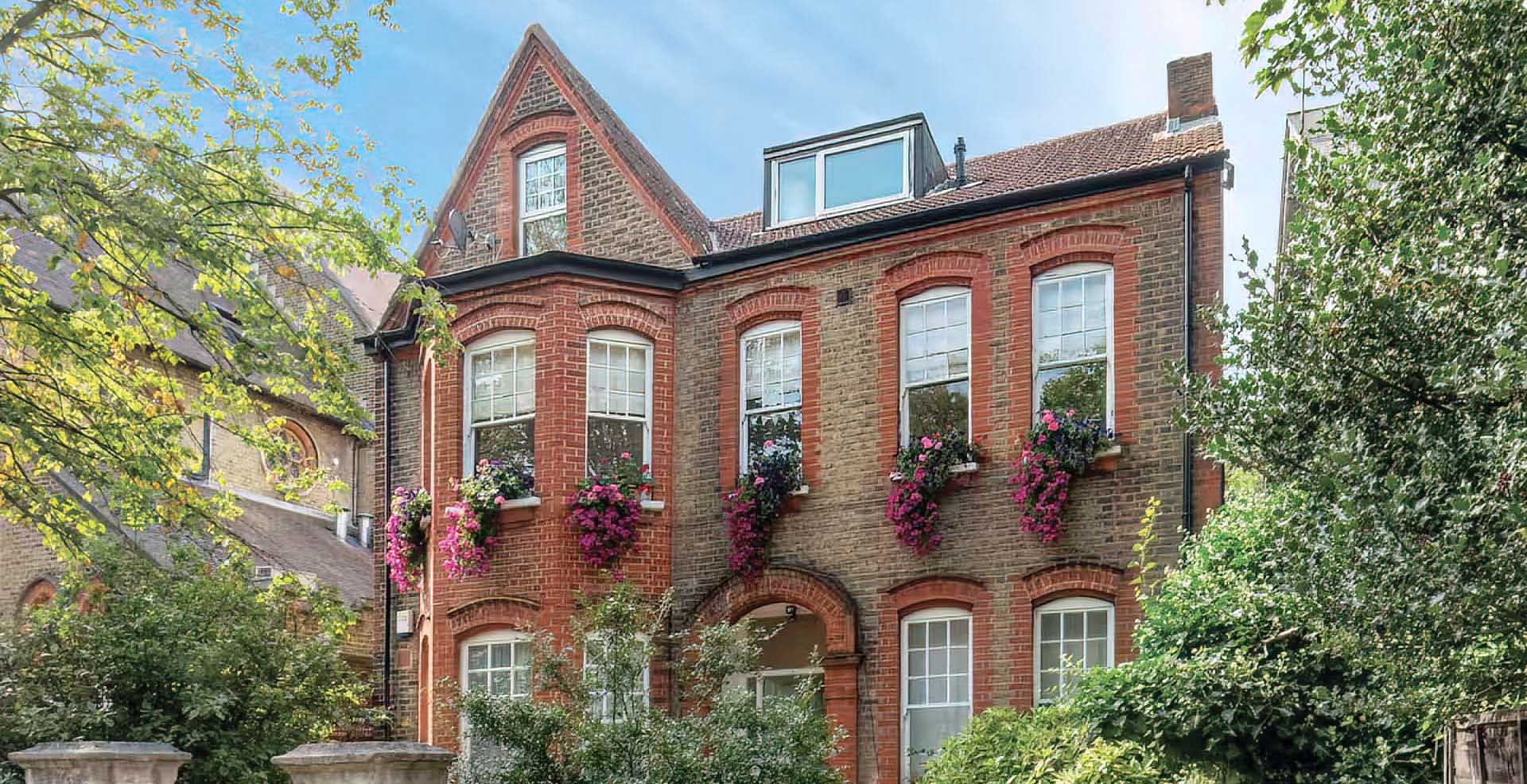 Historic brick building with arched windows adorned with pink and purple flowers, surrounded by lush trees, under a clear blue sky.