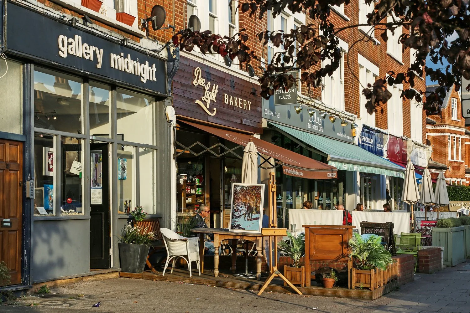Street view showing cafes and shops with outdoor seating, including 'gallery midnight,' 'Dev Light Bakery,' and 'Frelich Cafe,' with people sitting outside dining and artwork displayed on an easel.