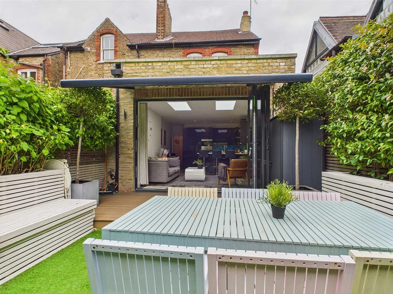 View of a modern backyard with outdoor table, benches, potted plants, and open sliding doors leading into a living room.