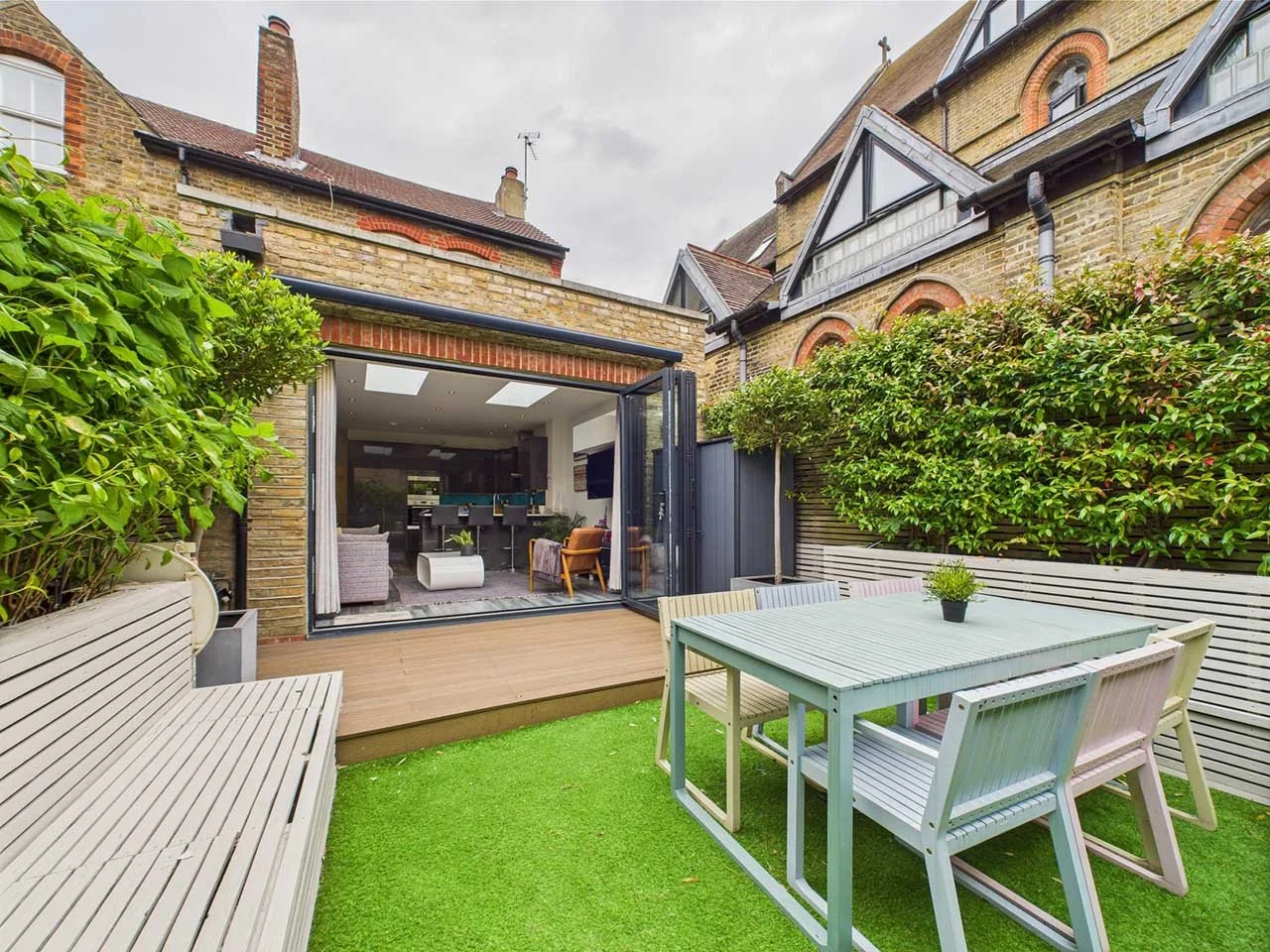Outdoor patio with table and chairs, adjacent to a modern interior living space, surrounded by greenery and enclosed by fences.
