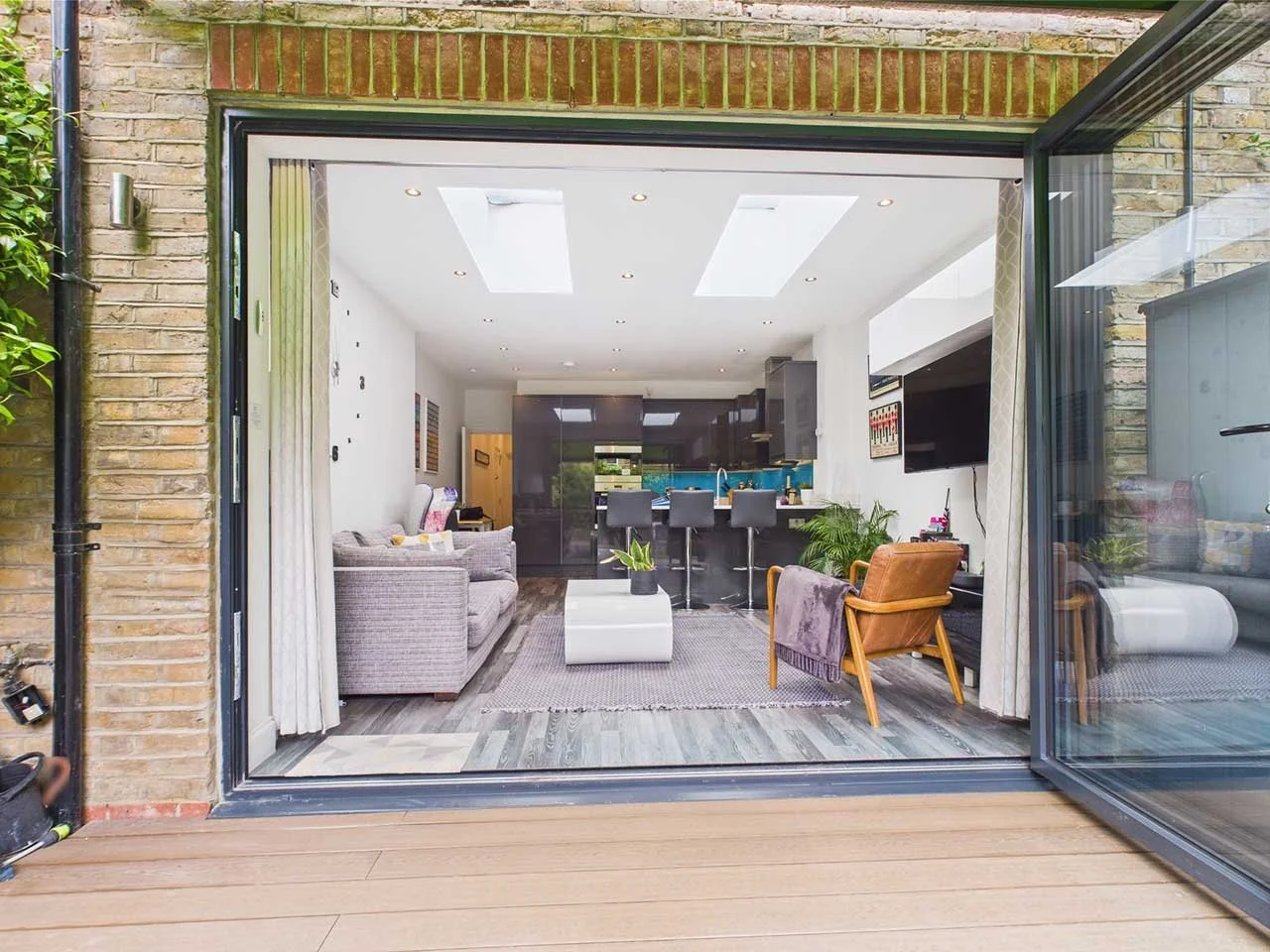 Open sliding glass door reveals a modern living room with a gray couch, an orange armchair with a purple blanket, a white coffee table with plants, a gray rug, and a kitchen in the background with bar stools, black cabinets, and skylights on the ceil