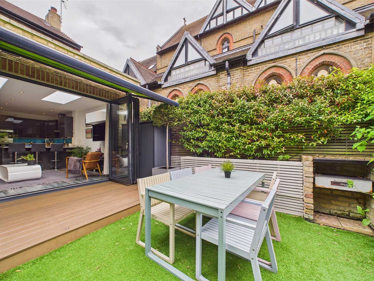 View of a backyard patio with outdoor dining table and chairs, adjacent to an open living room with a kitchen in the background. The space features a wooden deck, greenery, and a neighboring brick building with dormer windows.