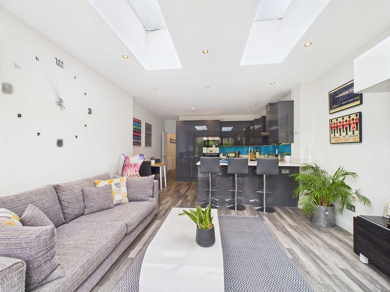 Modern open-plan living room with a gray sofa, a white coffee table with a potted plant, large windows on the ceiling, and a kitchen with gray cabinets, a breakfast bar, and four gray bar stools.