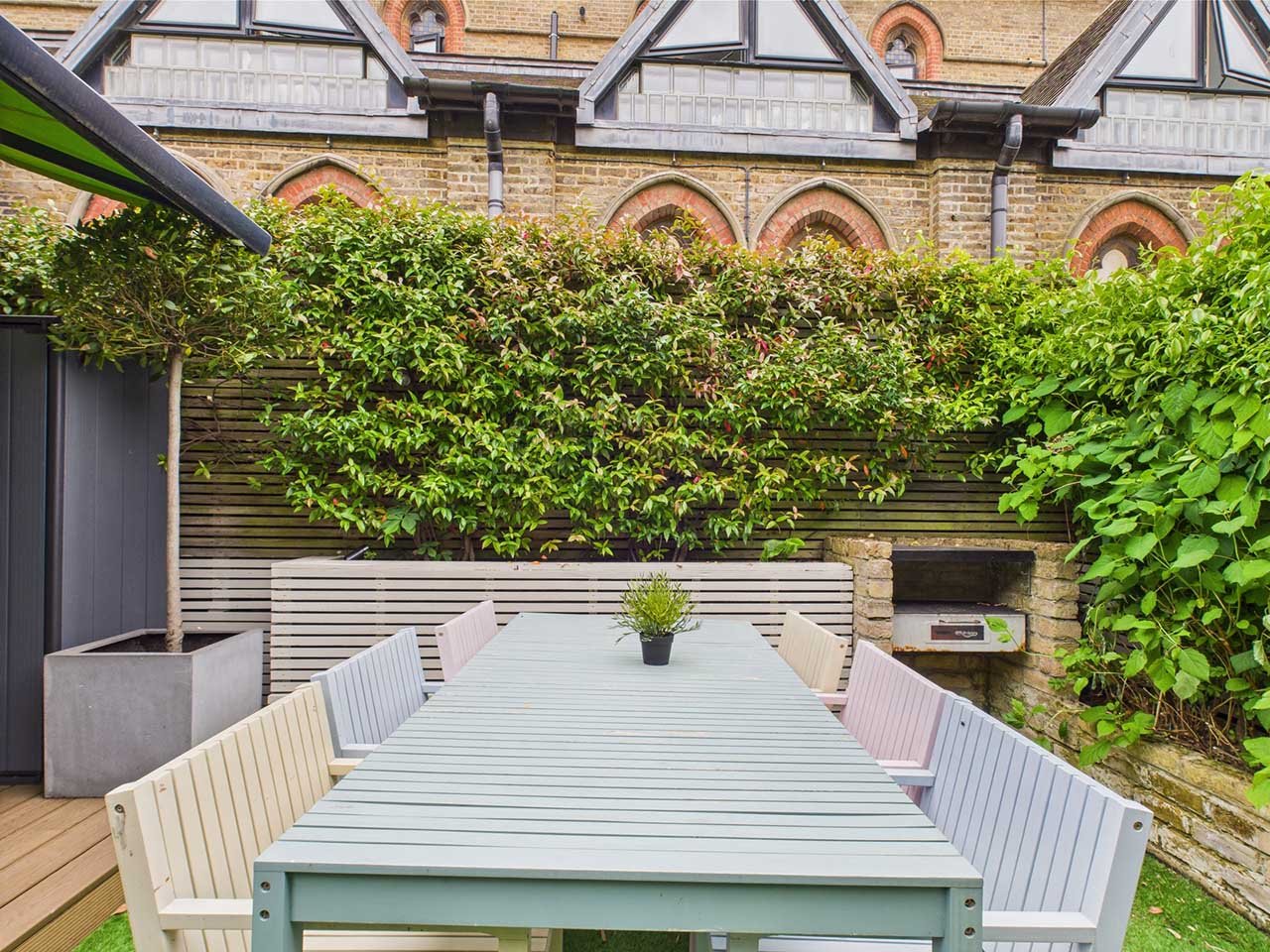 Outdoor dining area with a long table and six chairs, a potted plant in the center, surrounded by green bushes and a privacy screen, with an old brick building in the background.