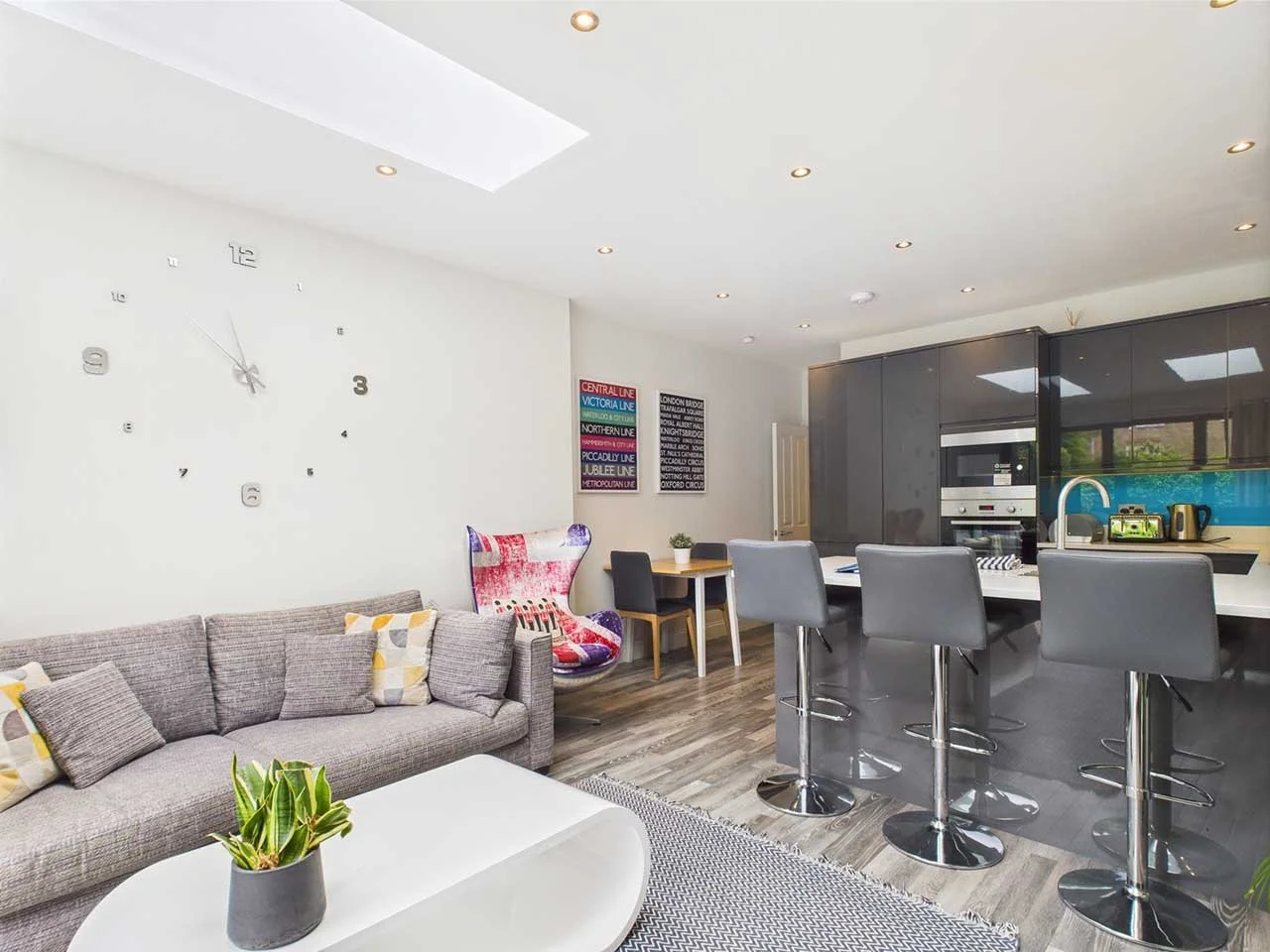 Modern living room and kitchen area with gray sofa, colorful artwork, wall clock, bar stools, and kitchen appliances.