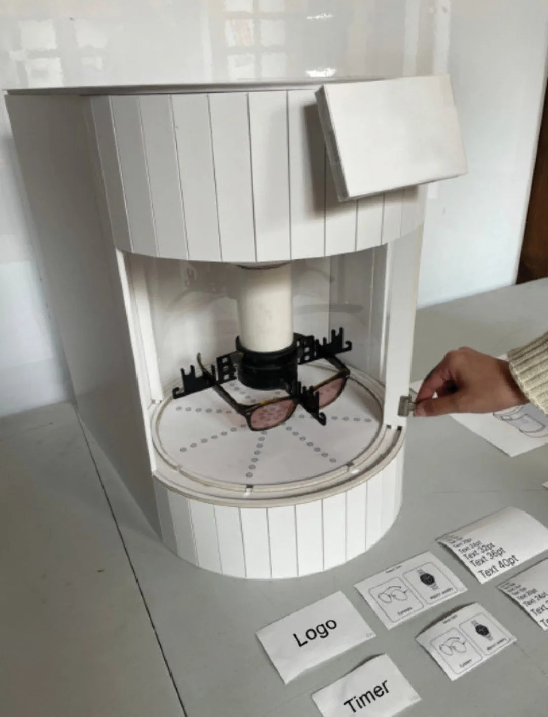 A white coffee vending machine with a slot for inserting coins or bills, partially disassembled, with switches and printed labels "Logo" and "Timer" nearby on a table.