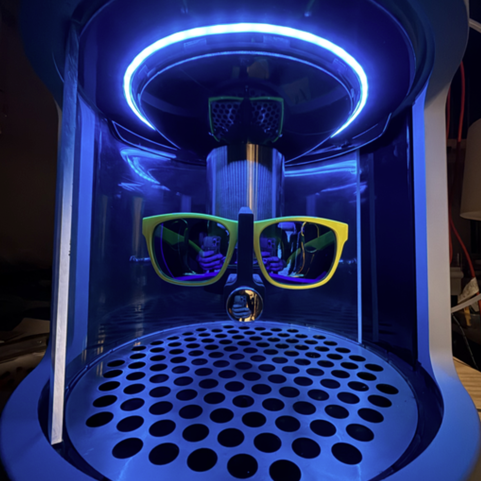 A black 3D printer with blue and green LED lights, with a pair of yellow glasses and a person taking a selfie reflected in the glass of the printer.